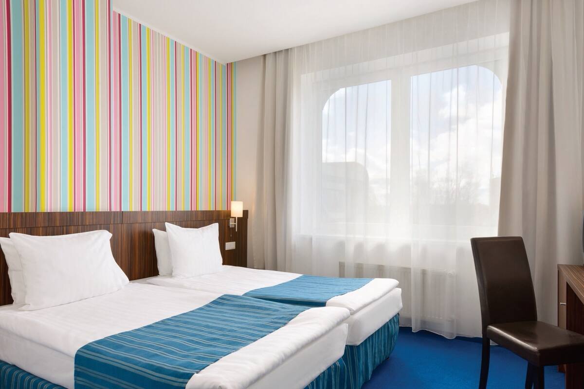 Ein Hotelzimmer mit einem großen Bett, einem Schreibtisch, einem Stuhl und einem Fenster mit durchscheinenden Vorhängen.
