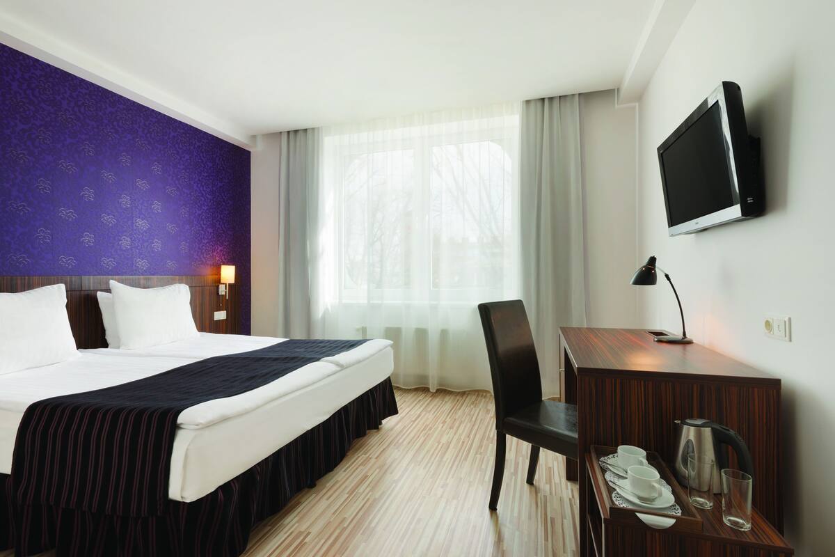 Ein Hotelzimmer mit einem Bett, einem Schreibtisch mit Stuhl, einem Fernseher und einem Fenster mit Vorhängen.