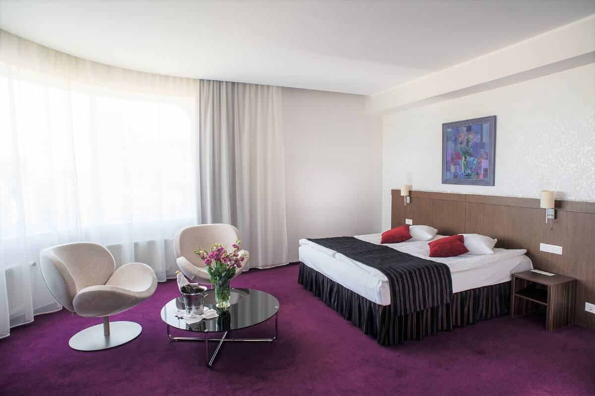 Ein Hotelzimmer mit einem Bett, zwei Sesseln, einem kleinen Tisch mit Blumenvase und einem Gemälde an der Wand.
