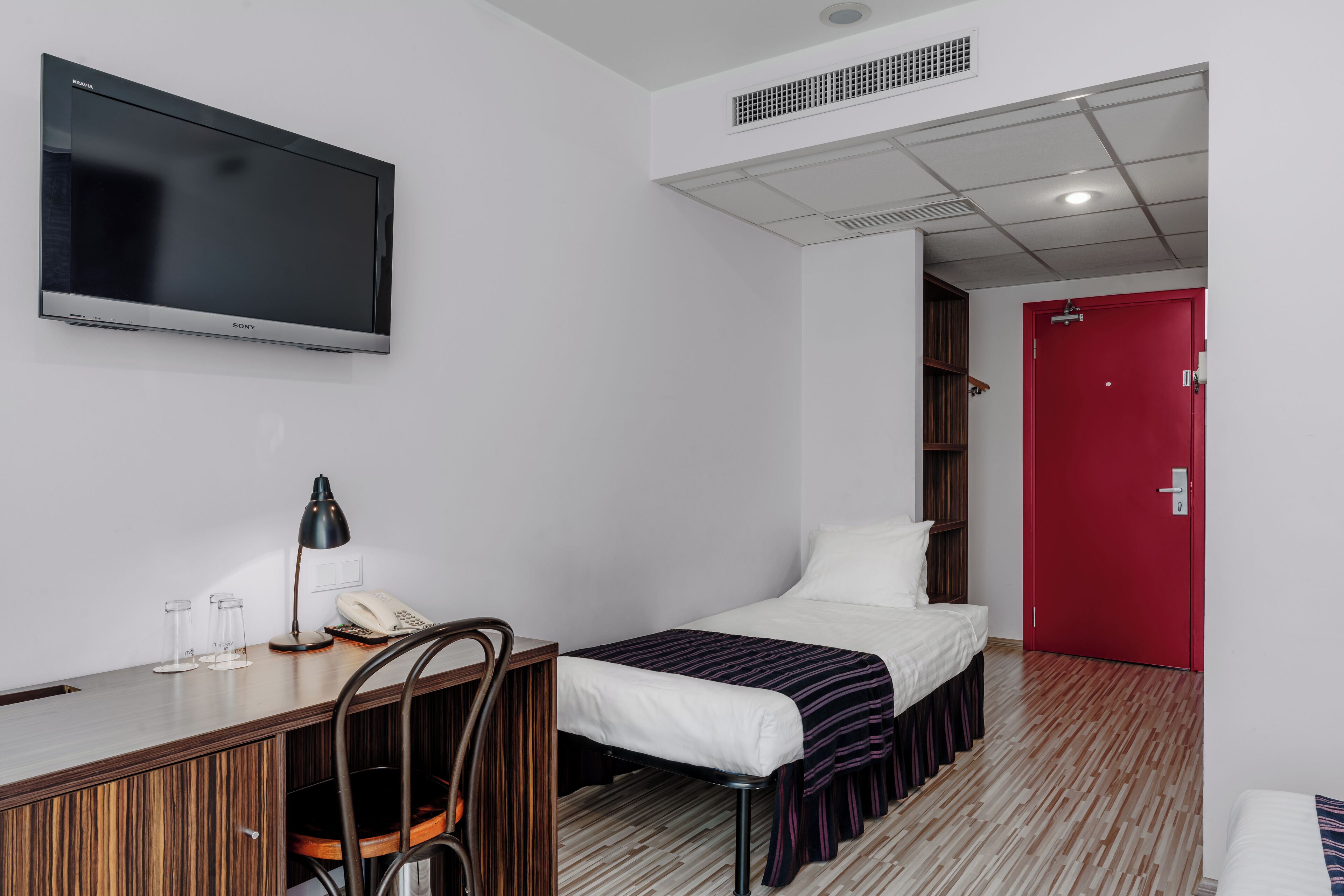 A hotel room with a bed, a desk with a chair, a television, and a red door.
