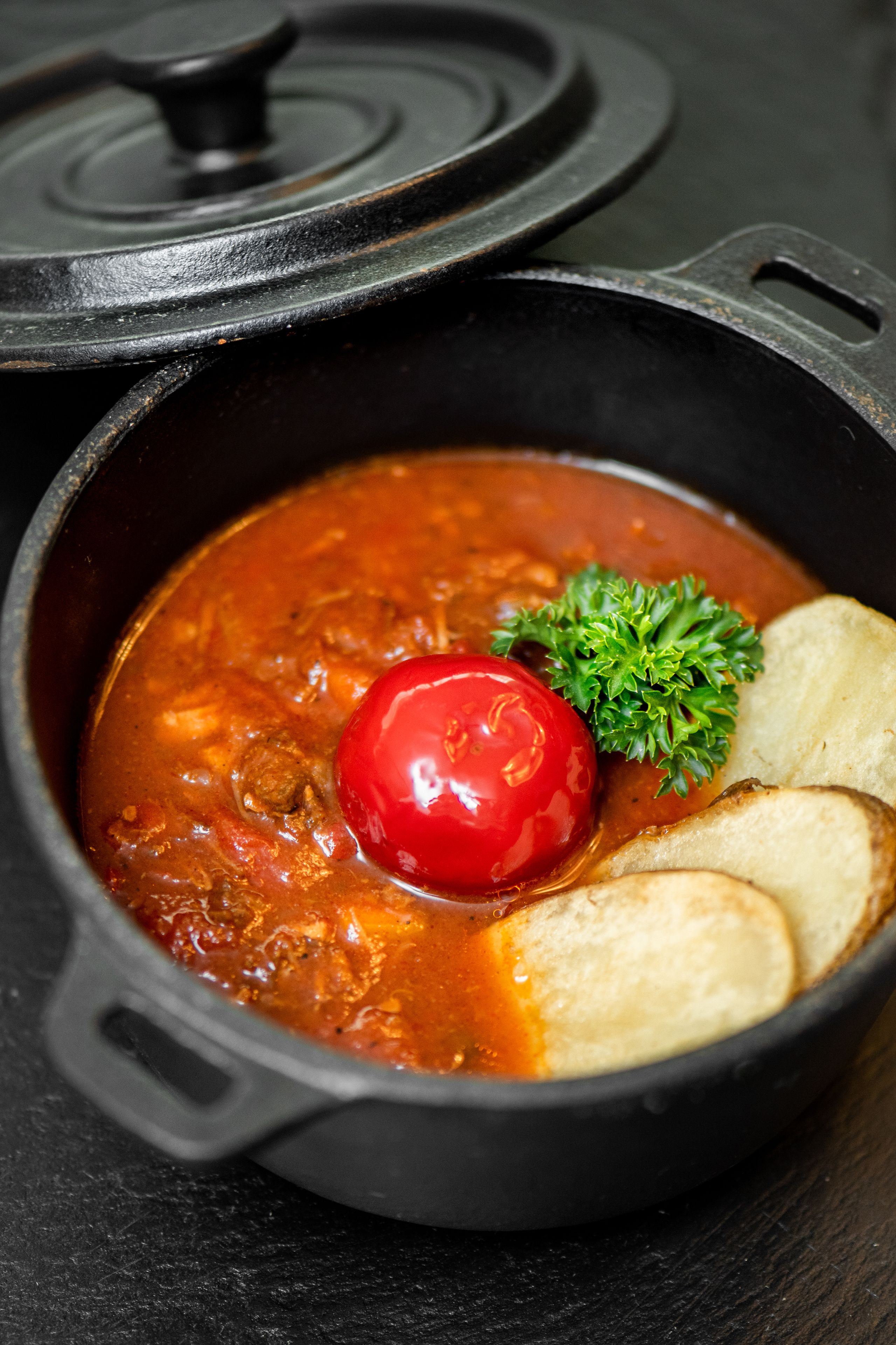 Un guiso en una olla con tapa, un tomate y pan sobre una superficie oscura.