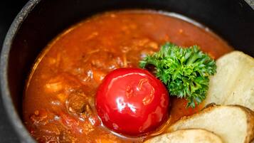 Un guiso en una olla con tapa, un tomate y pan sobre una superficie oscura.