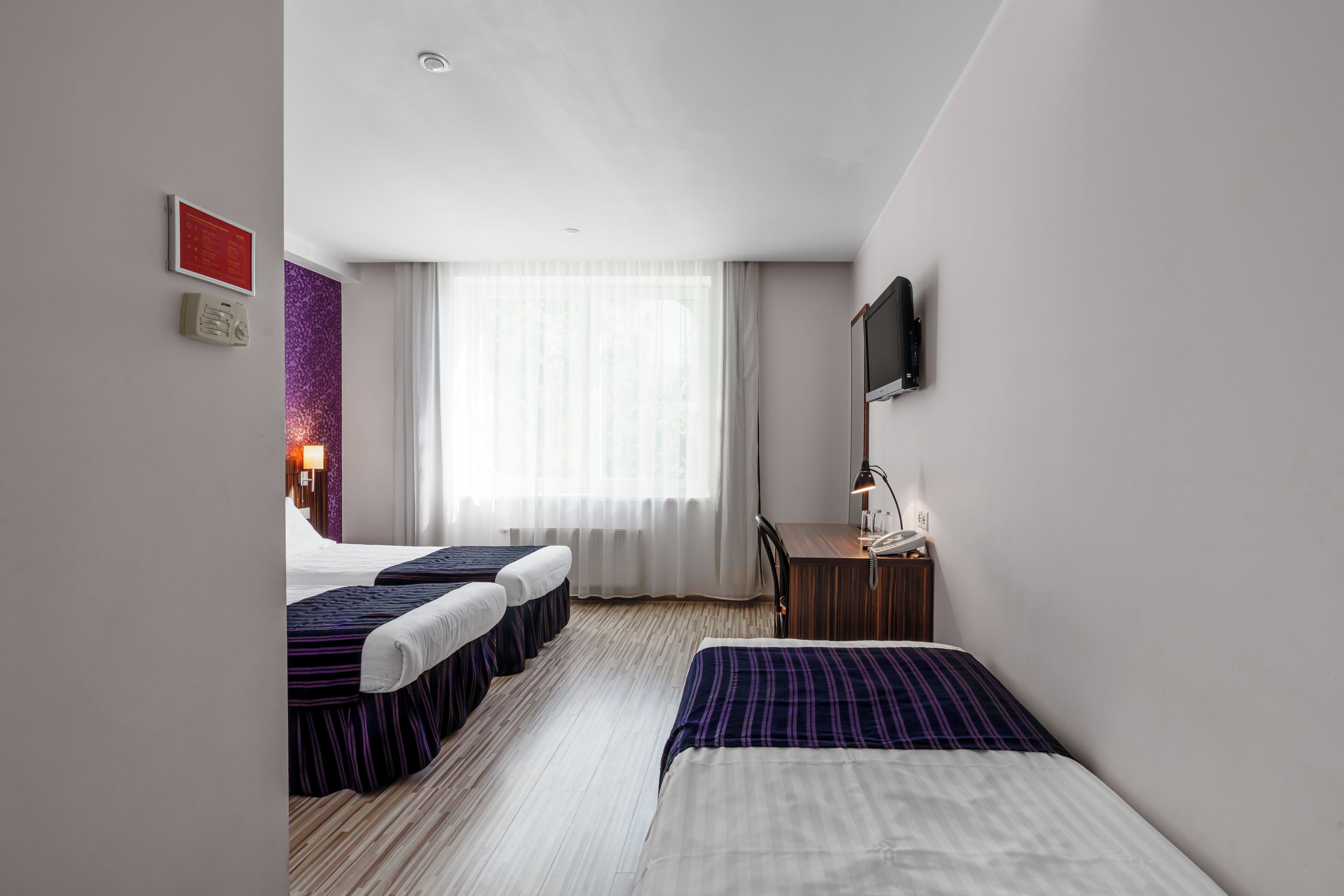 A hotel room with two beds, a desk, a TV, and a window with sheer curtains.