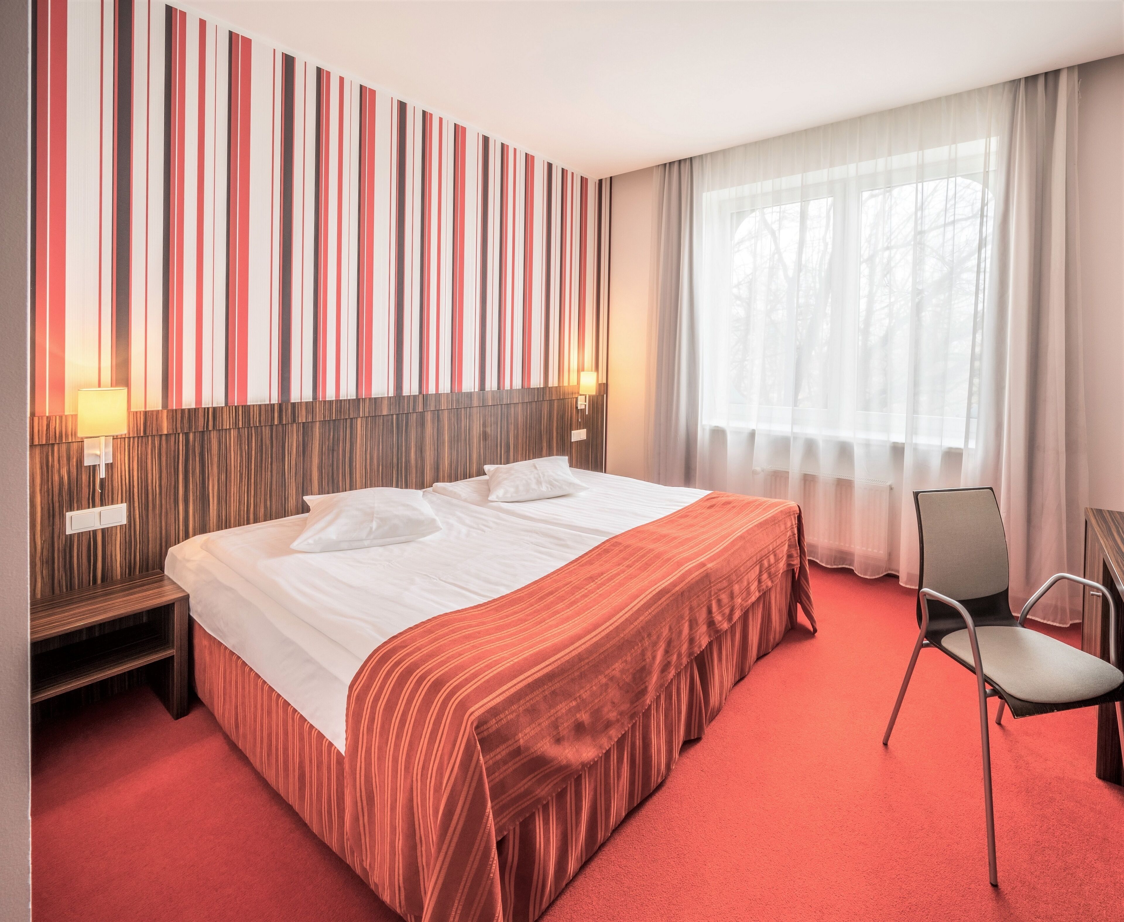 Hotellihuone, jossa on suuri sänky, puinen pääty, punainen matto, tuoli ja ikkuna, jossa on läpikuultavat verhot.
