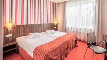 Une chambre d’hôtel avec un grand lit, une tête de lit en bois, une moquette rouge, une chaise et une fenêtre ornée de rideaux légers.