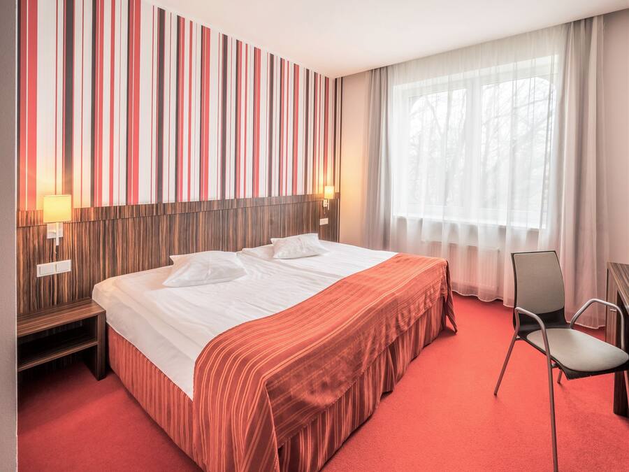 Une chambre d’hôtel avec un grand lit, une tête de lit en bois, une moquette rouge, une chaise et une fenêtre ornée de rideaux légers.