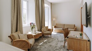 Et hotelværelse med sofa, to lænestole, et lille bord med blomster i en vase, en lampe og et fladskærms-tv på væggen.