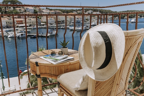 A wicker chair with a straw hat, a small table with a plant, and a view of a marina with boats.
