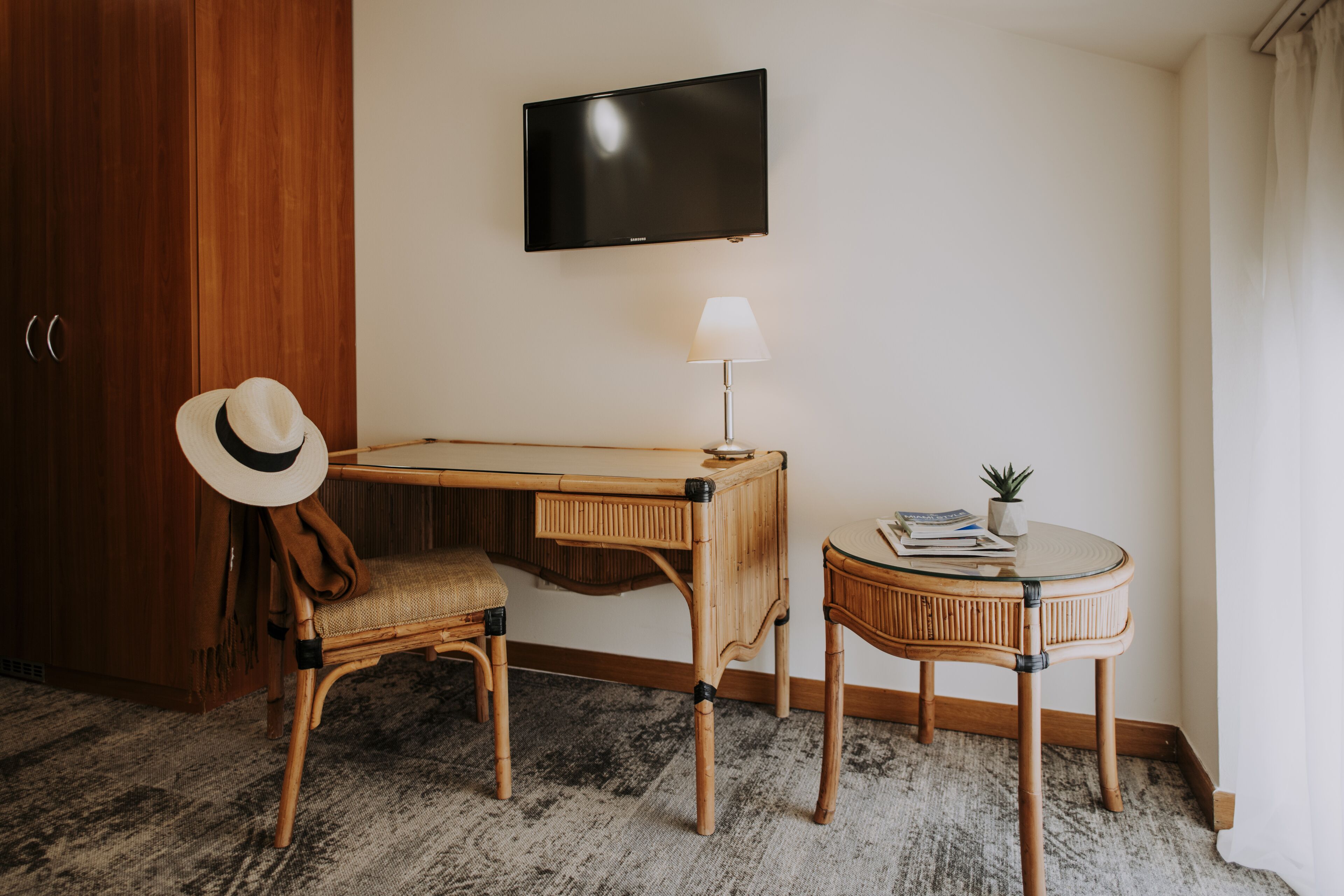 A room with a wooden desk, a chair, a small round table with a lamp, a television on the wall, and a hat on the chair.