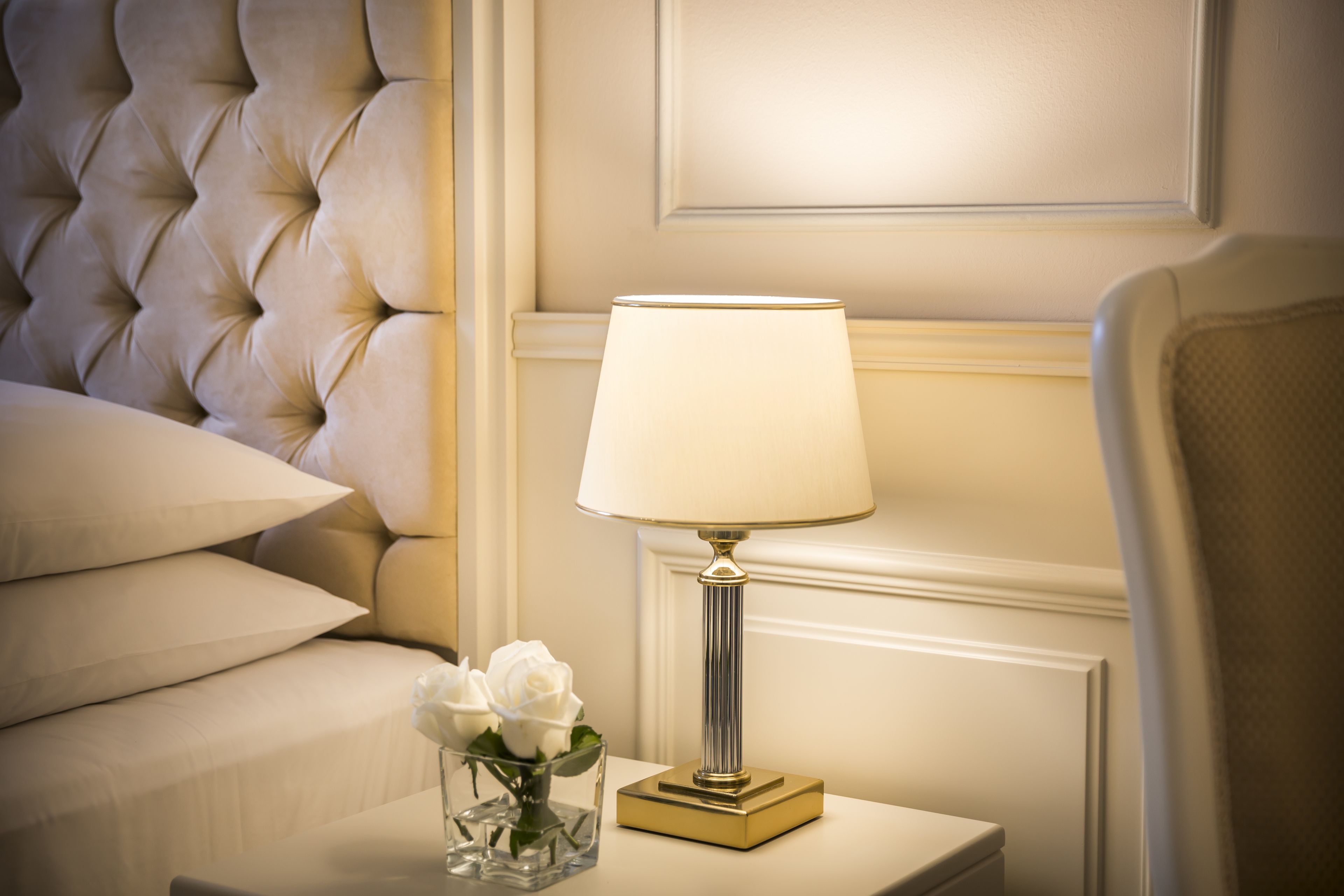 A neatly arranged bedroom with a tufted headboard, a bedside table with a lamp, and a vase with white roses.