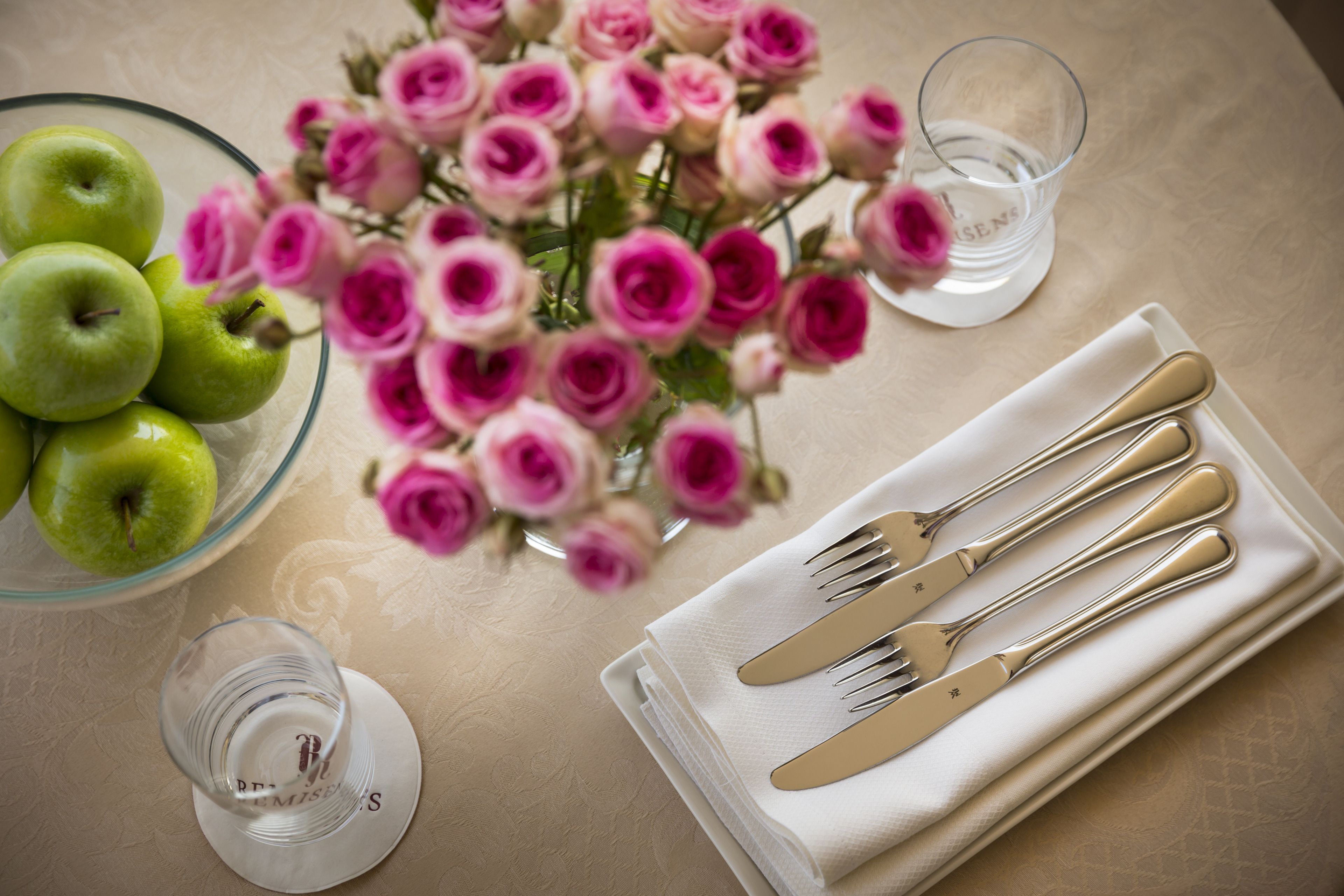 A table setting with a bowl of green apples, a vase of pink roses, and a glass of water.
