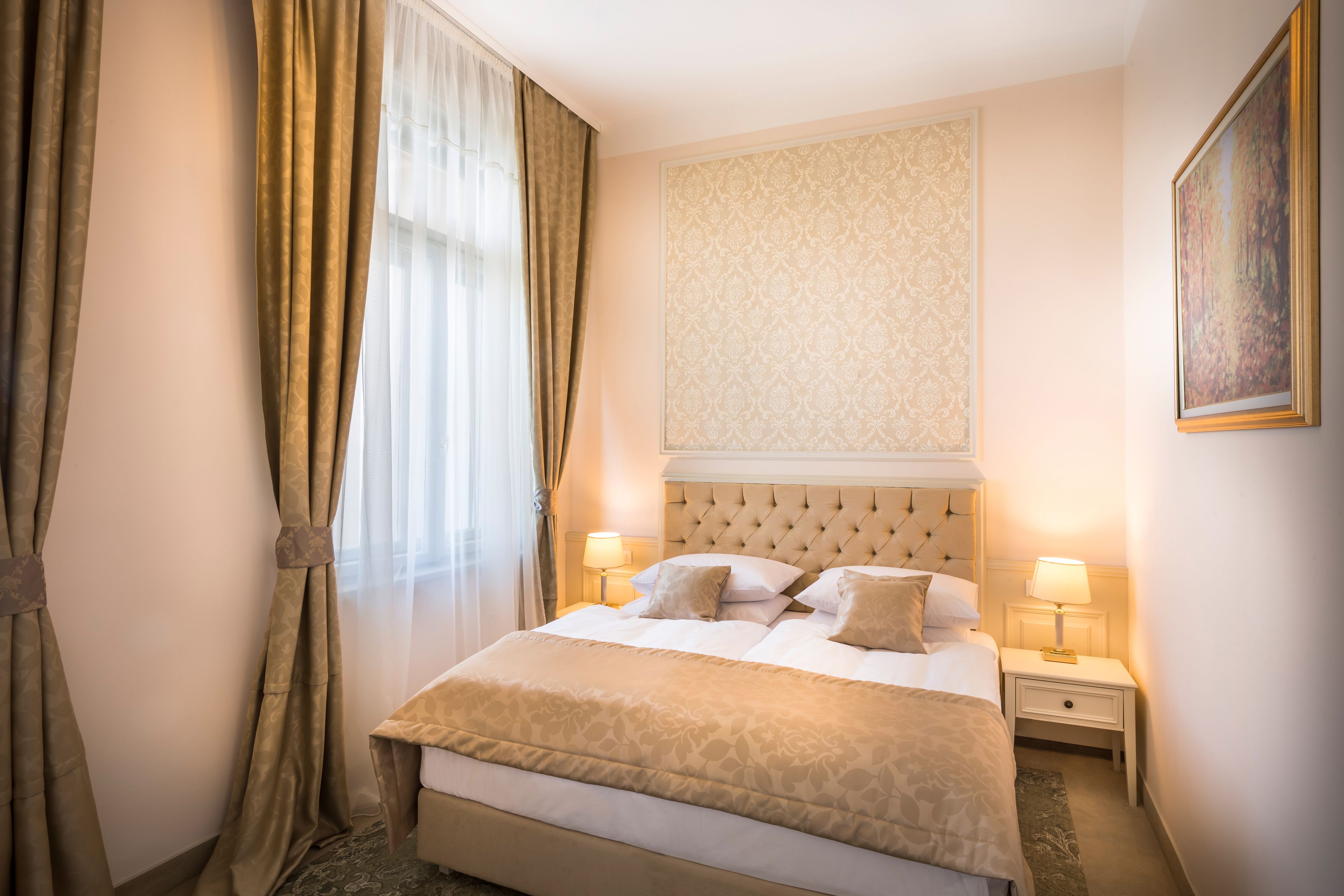 A hotel room with a large bed, two bedside tables with lamps, a decorative headboard, and a painting on the wall.