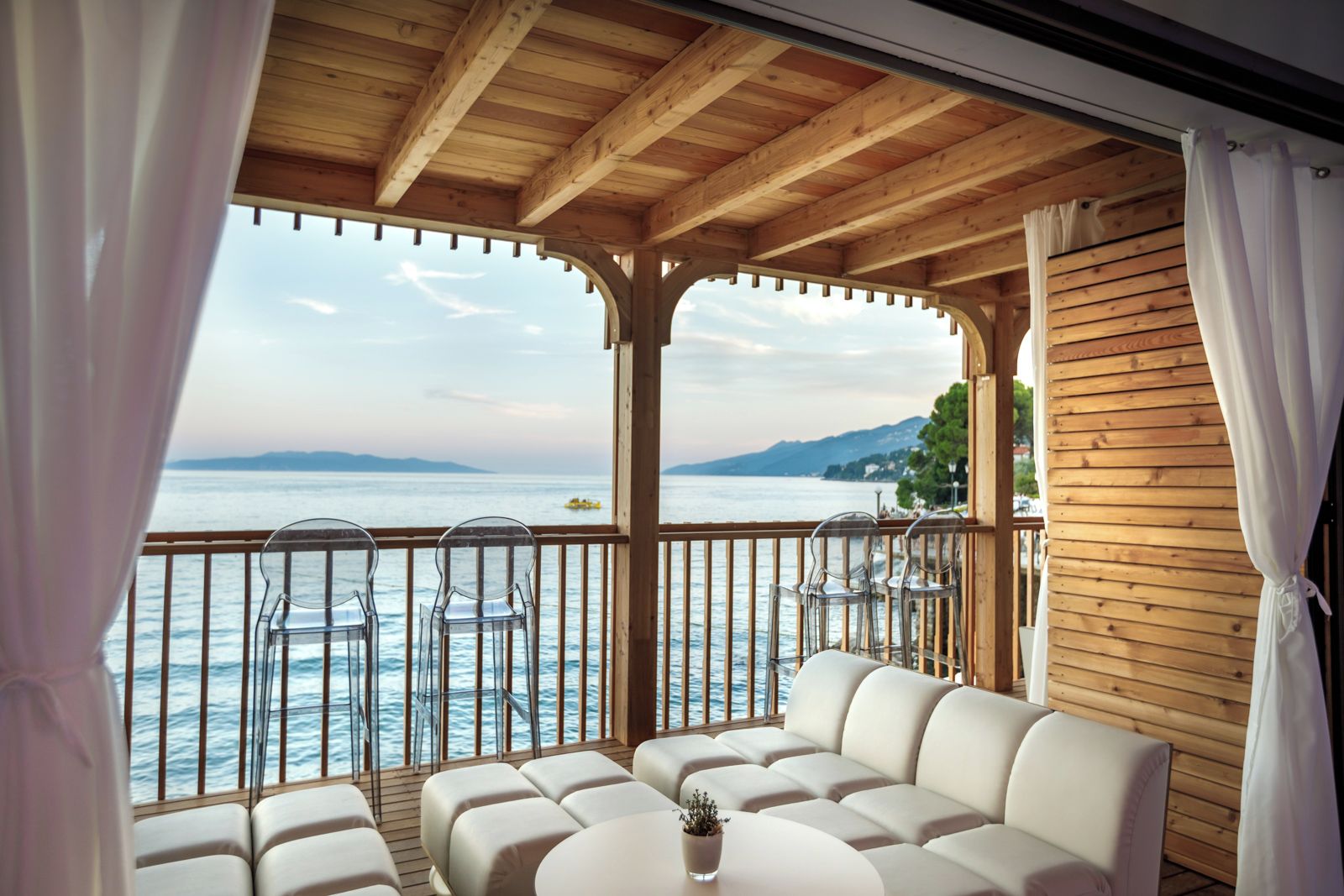 A covered outdoor seating area with a view of the sea, wooden beams, and clear chairs.
