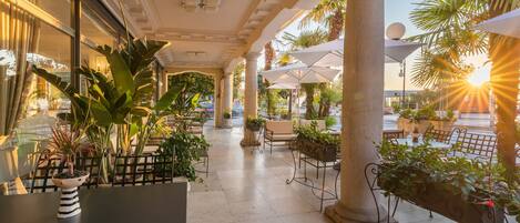 An outdoor patio area with tables and chairs, surrounded by columns and potted plants.