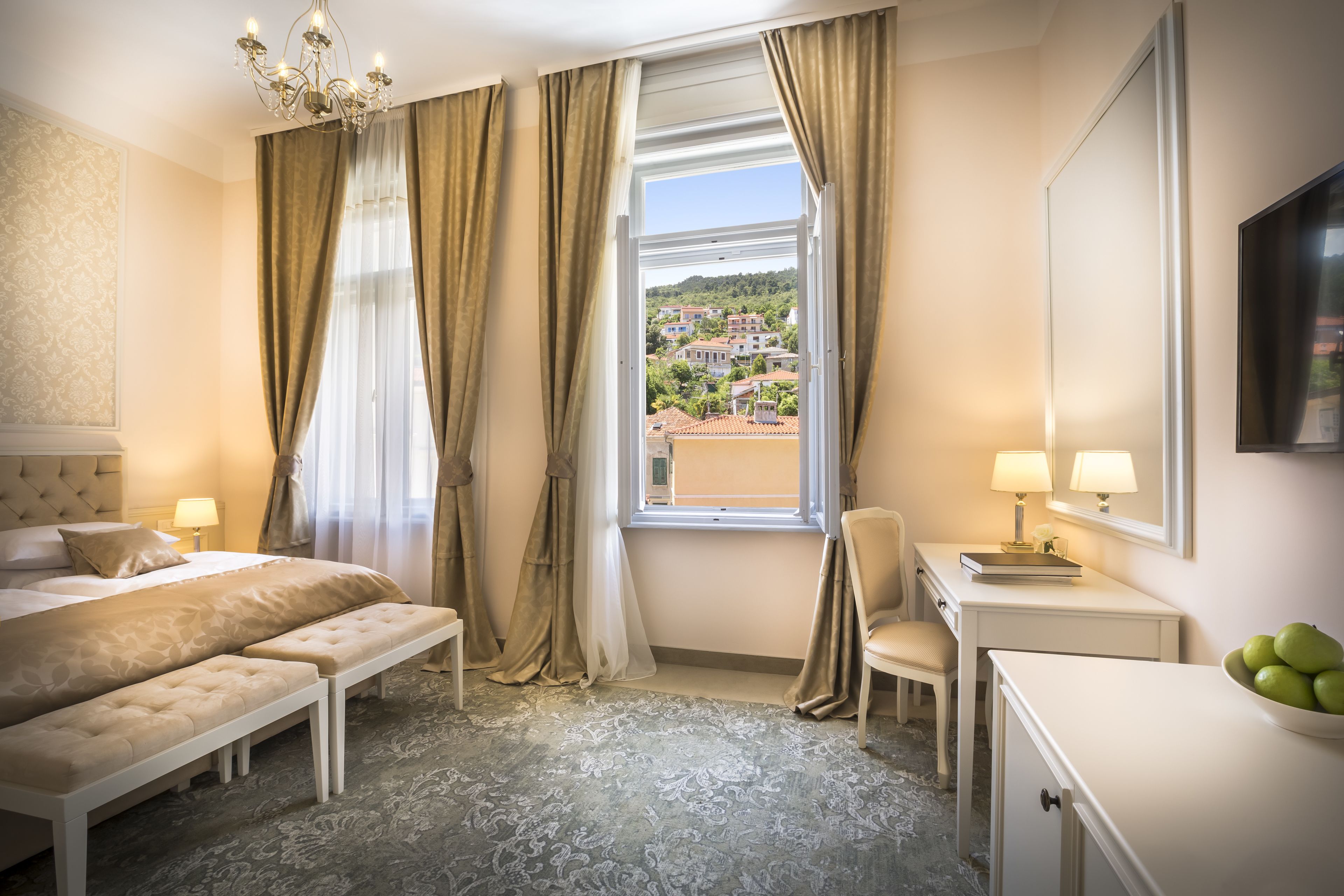 Habitación de hotel con cama, banco, escritorio, silla y vista a un pueblo a través de una ventana.