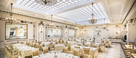 A spacious banquet hall with high ceilings, ornate chandeliers, and large mirrors. Tables are set for a formal event.