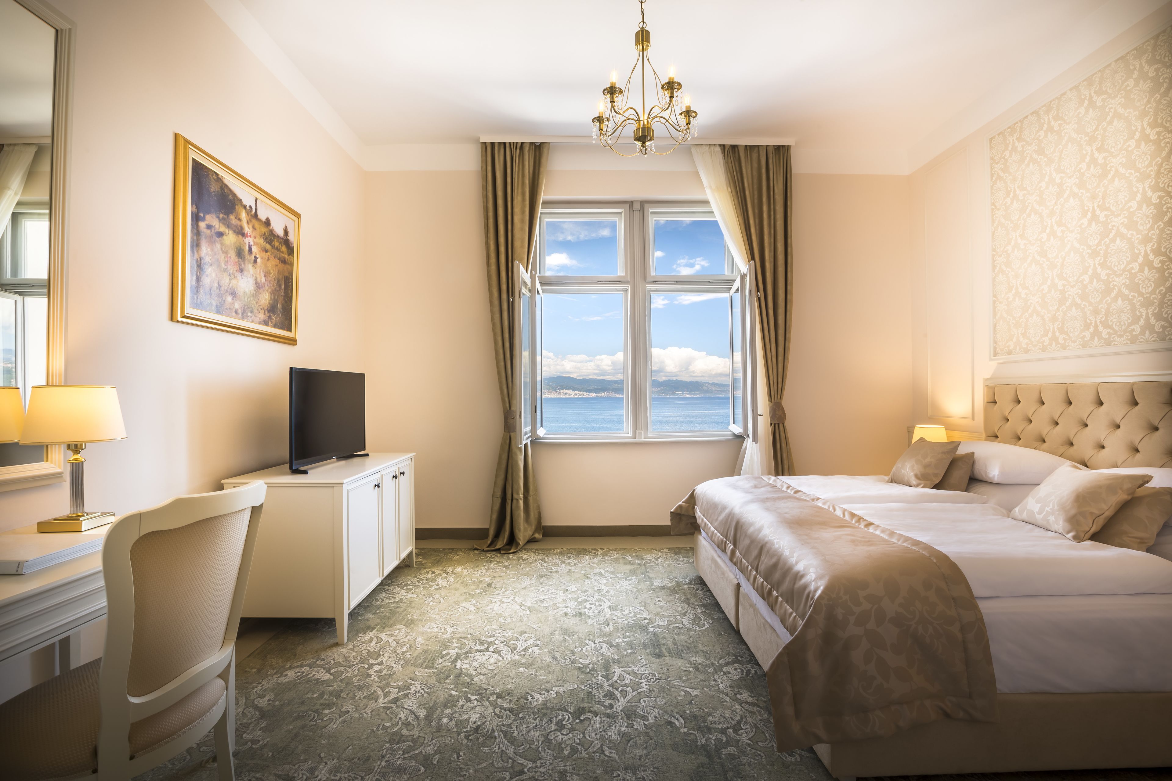 A hotel room with a large bed, a desk with a TV, a chair, and a view of the sea.