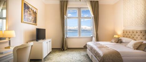 A hotel room with a large bed, a desk with a TV, a chair, and a view of the sea.