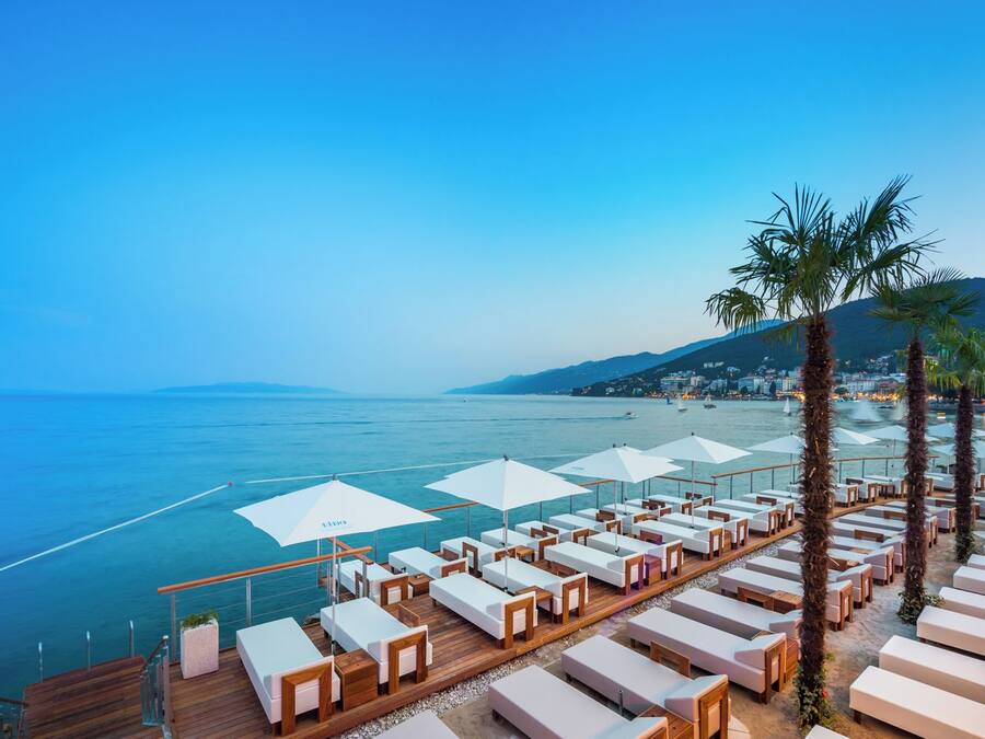 Ein Küstenort mit Liegestühlen und Sonnenschirmen, einer Holzterrasse und Blick auf das Meer und die entfernten Hügel.