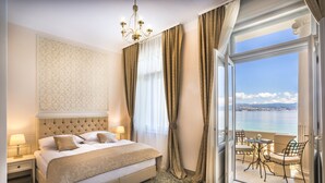 Een hotelkamer met een groot bed, een kroonluchter, een balkon met een tafel en stoelen en uitzicht op zee.