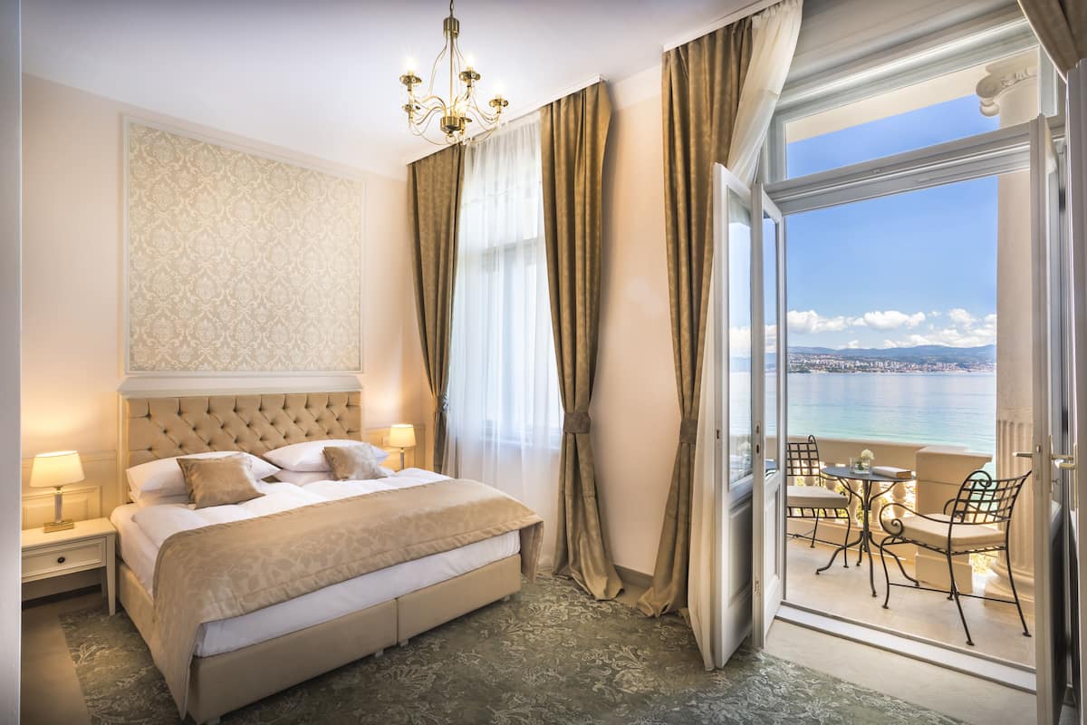 Ein Hotelzimmer mit einem großen Bett, einer Kristallleuchter, einem Balkon mit Tisch und Stühlen sowie Meerblick.