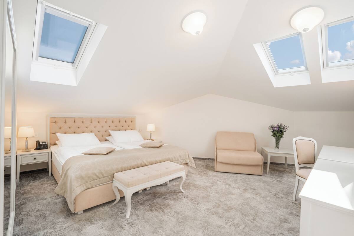 Ein Schlafzimmer mit einem großen Bett, einem Sessel, einem Schreibtisch und einem kleinen Tisch. Es gibt zwei Fenster mit Dachfenstern und eine Deckenleuchte.