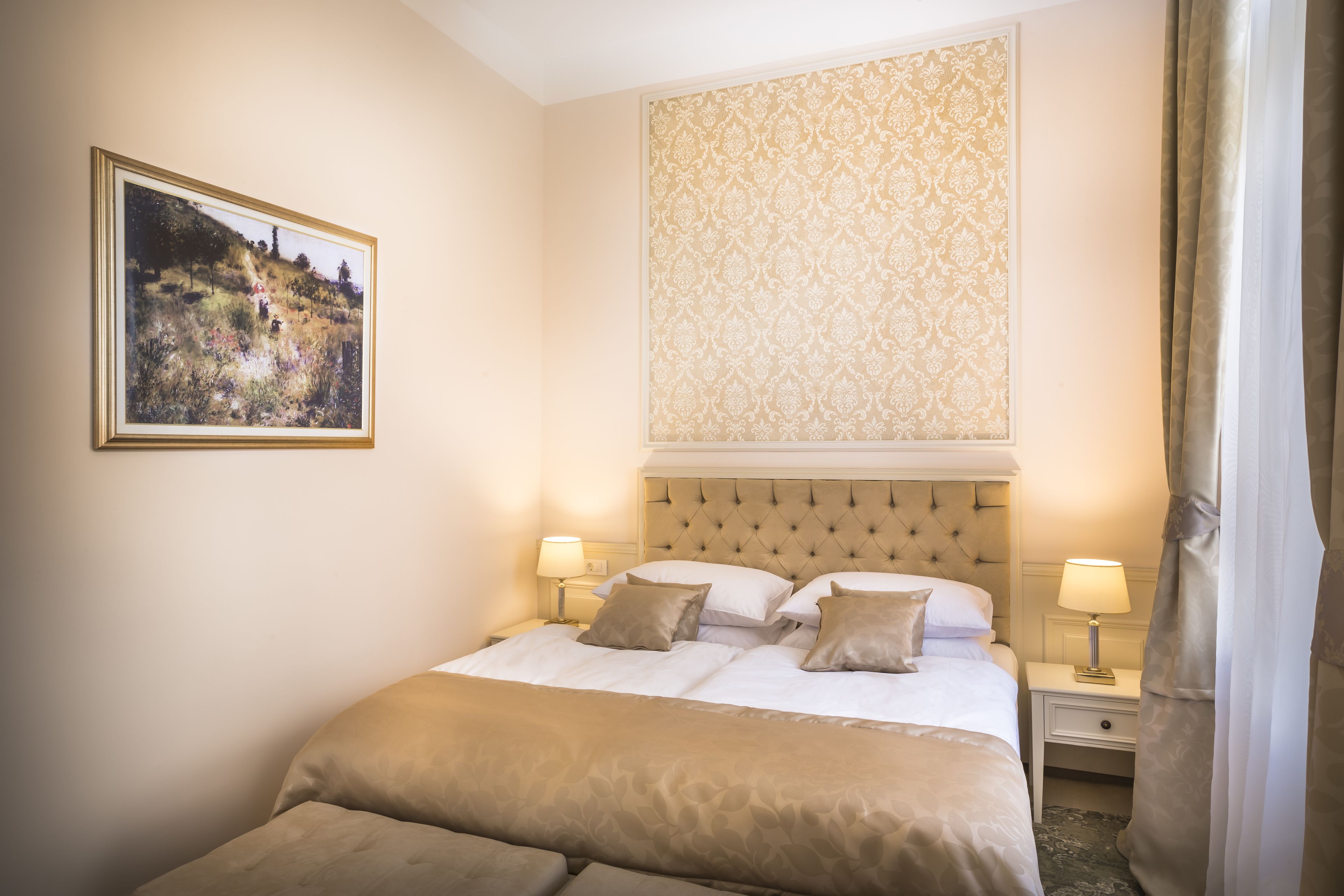 A neatly arranged hotel room with a large bed, two bedside tables with lamps, a framed picture on the wall, and a patterned wallpaper above the bed.