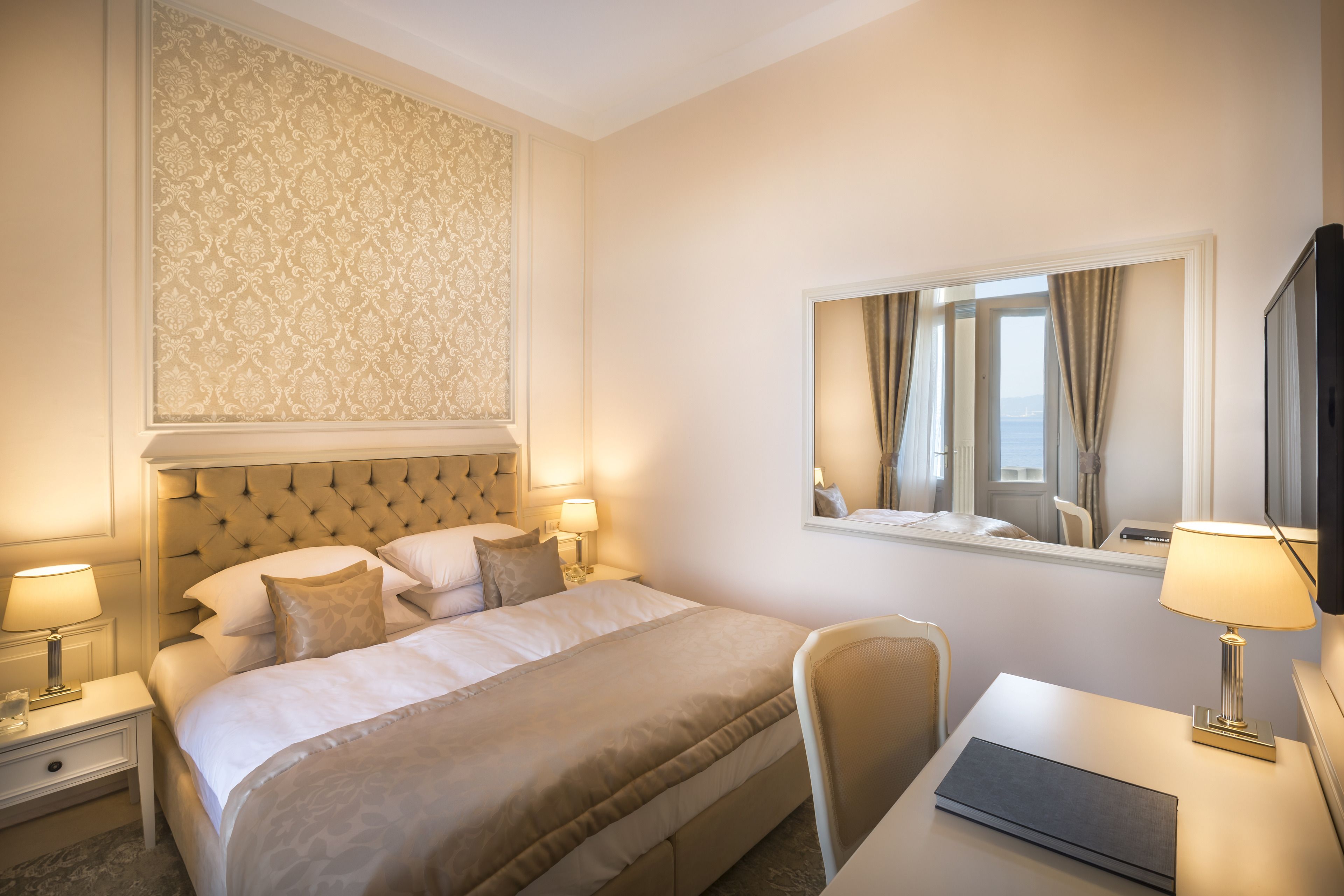 A hotel room with a large bed, a desk, a chair, a mirror, and a view of the sea through the window.