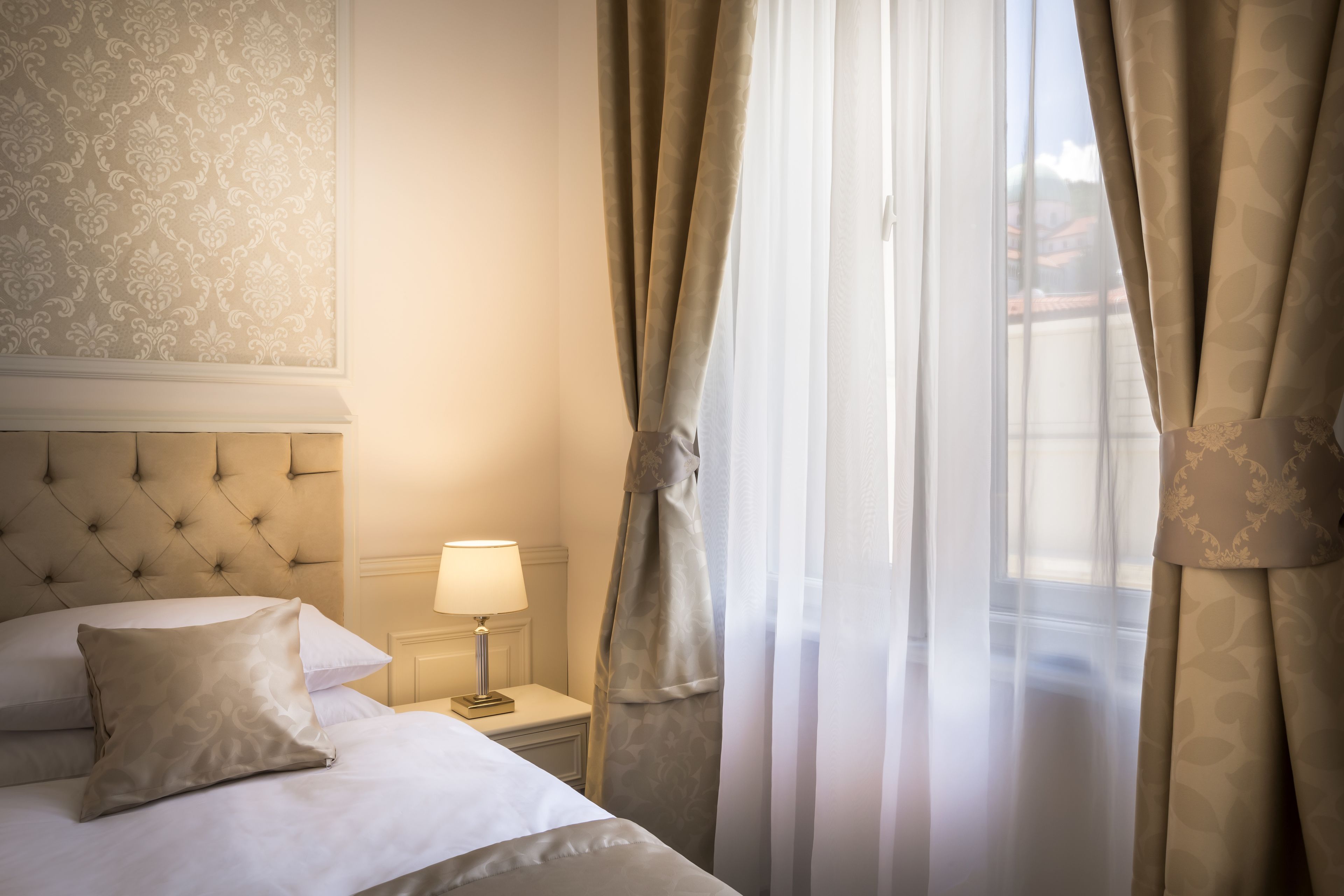 A hotel room with a tufted headboard, a bedside table with a lamp, and sheer curtains.
