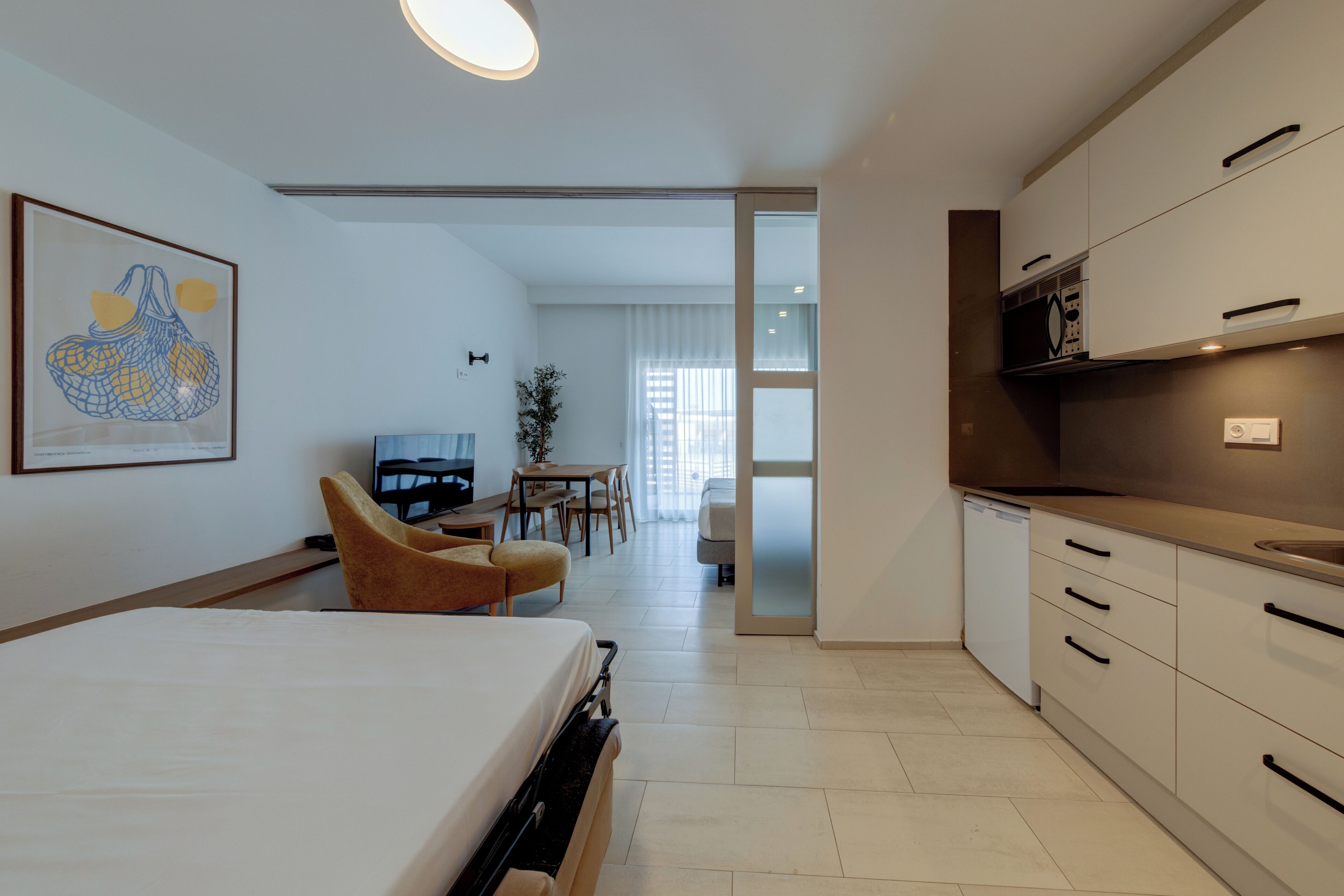 A modern hotel room with a bed, a desk, a chair, a TV, and a kitchen area.