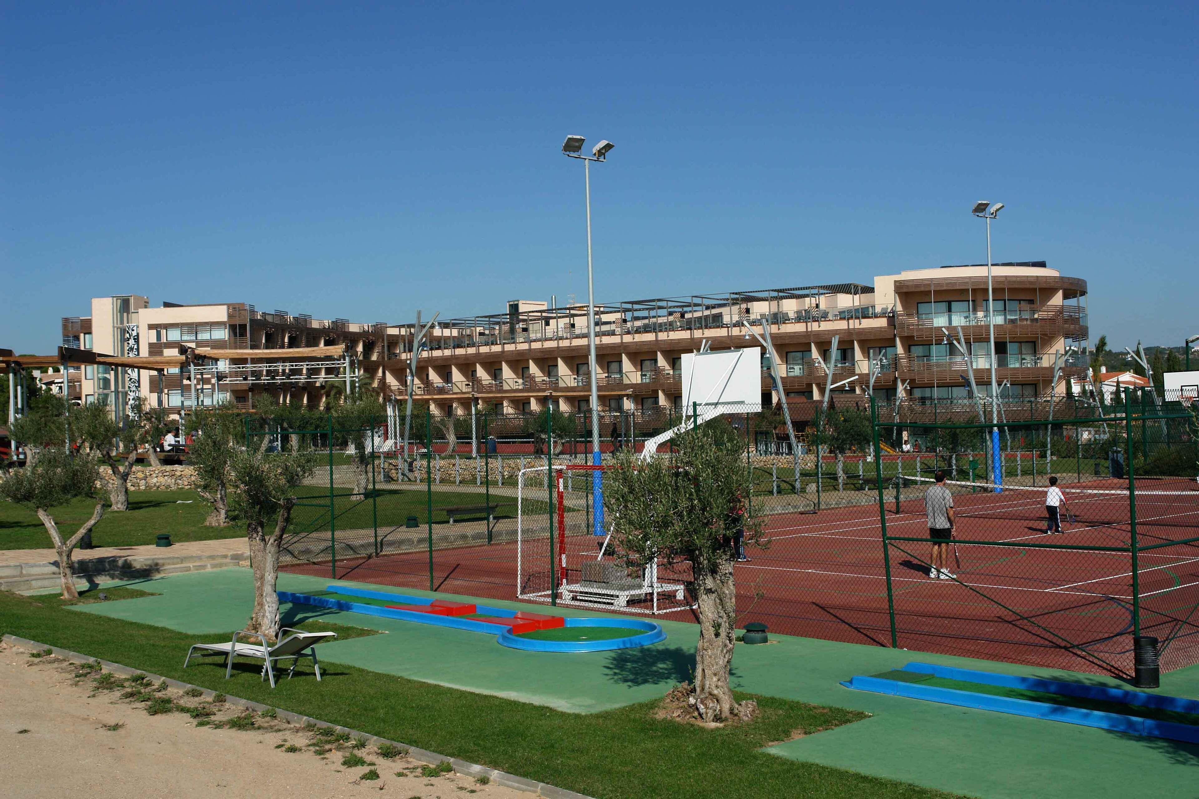 Un court de tennis avec un filet, entouré d’une clôture, et un bâtiment avec de nombreux balcons en arrière-plan.