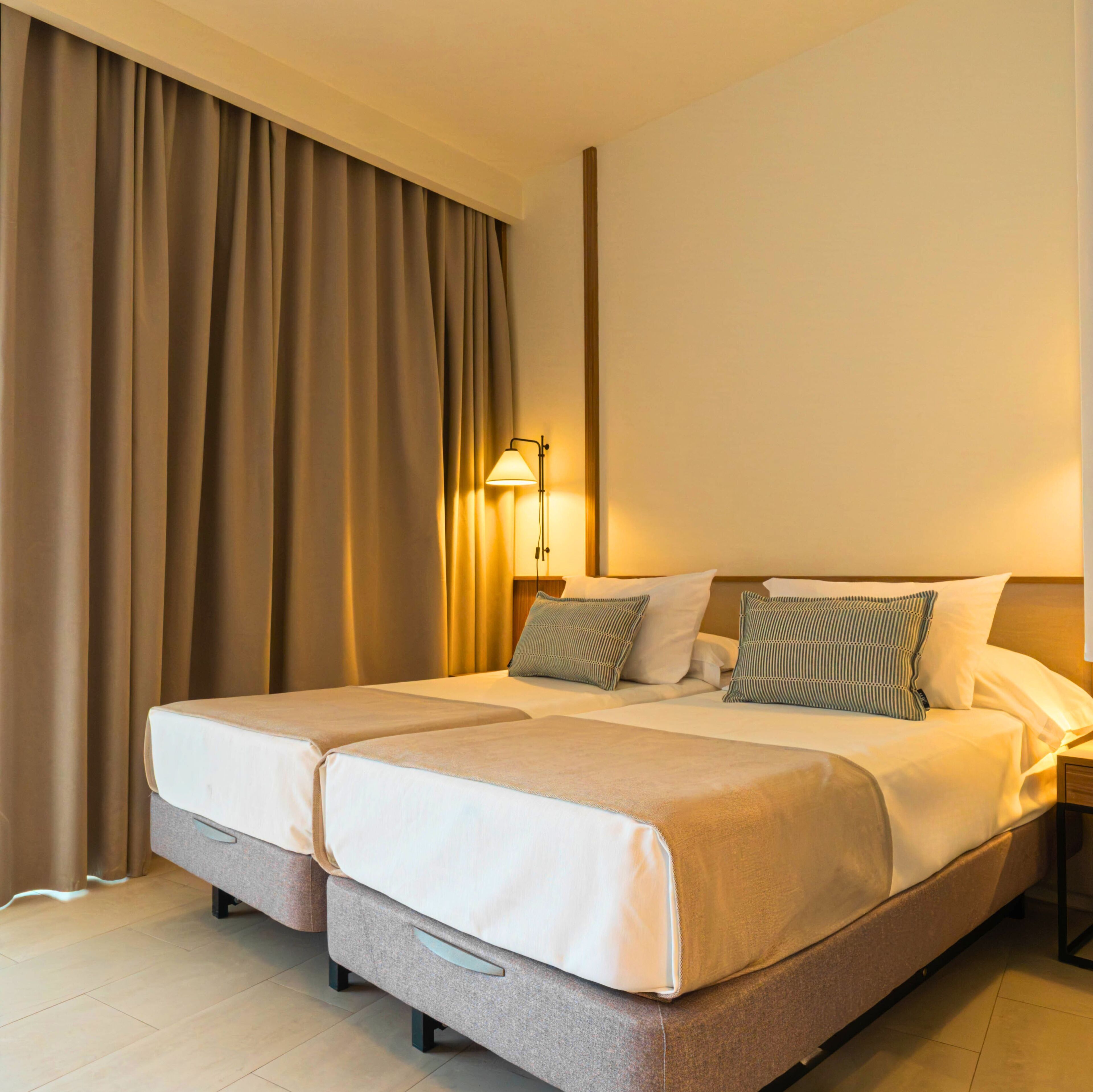 A hotel room with a double bed, two bedside tables, a lamp, and a large window with curtains.