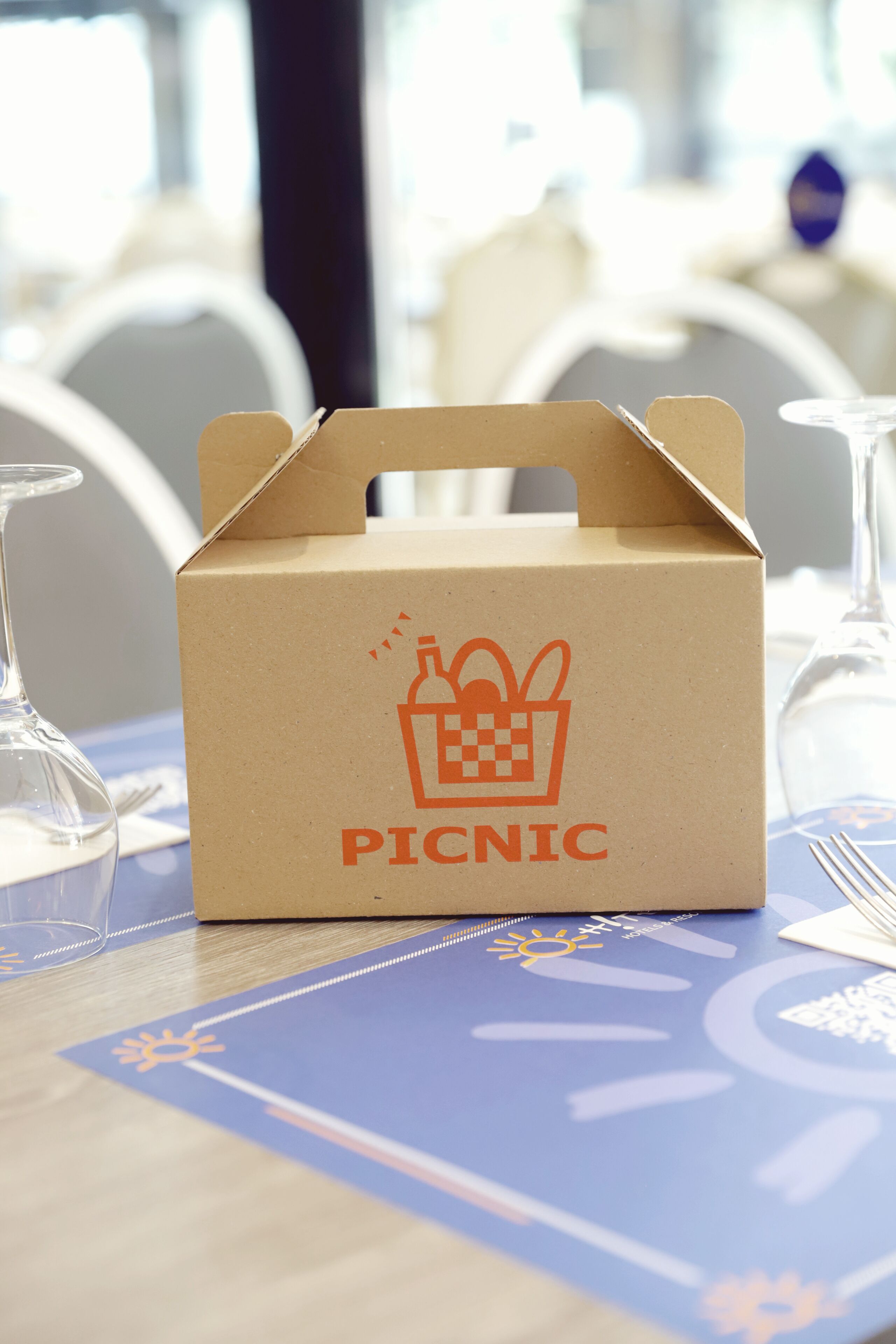 Une boîte à pique-nique en carton, ornée d’une icône représentant un panier et une bouteille, et sur laquelle est inscrit le mot « PIQUE-NIQUE », est posée sur une table recouverte d’une nappe bleue, entourée de verres à vin.