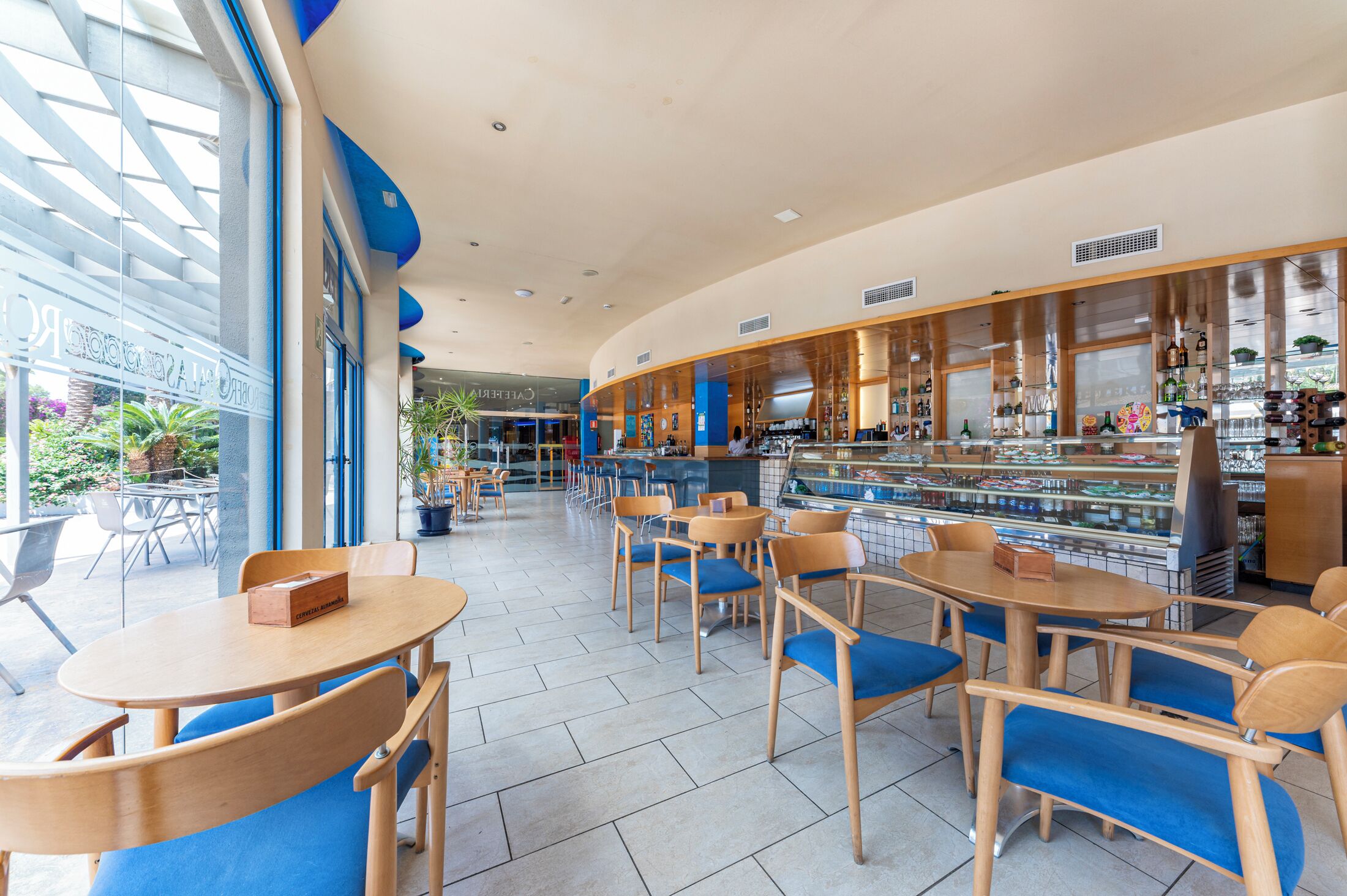 Una cafetería moderna con mesas de madera y sillas azules, una zona de bar con vitrinas de vidrio y grandes ventanales con vistas al exterior.