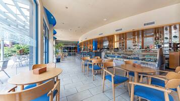 Una cafetería moderna con mesas de madera y sillas azules, una zona de bar con vitrinas de vidrio y grandes ventanales con vistas al exterior.