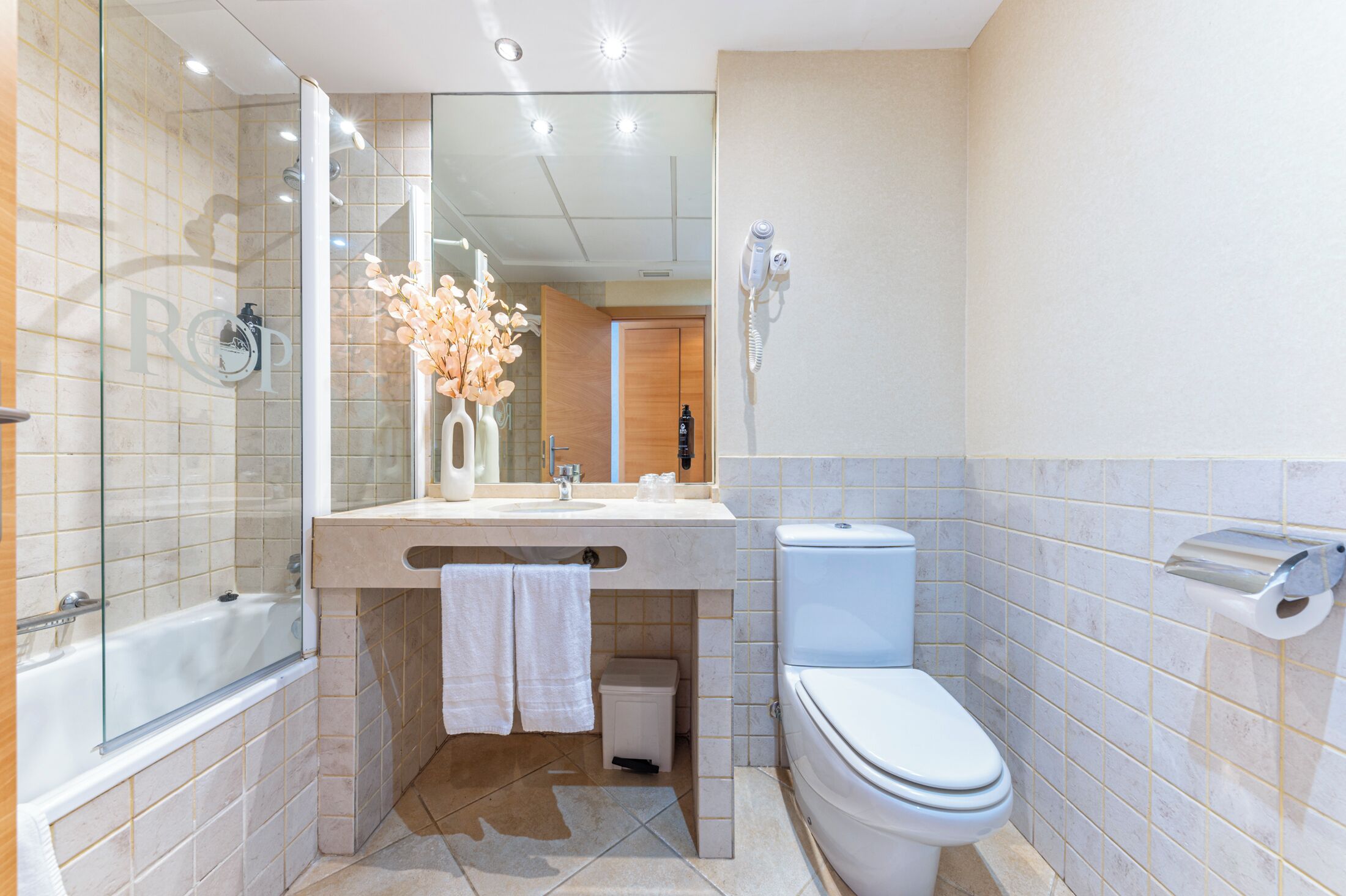 A modern bathroom with a glass-enclosed shower, a vanity with a mirror, a toilet, and a hairdryer mounted on the wall.