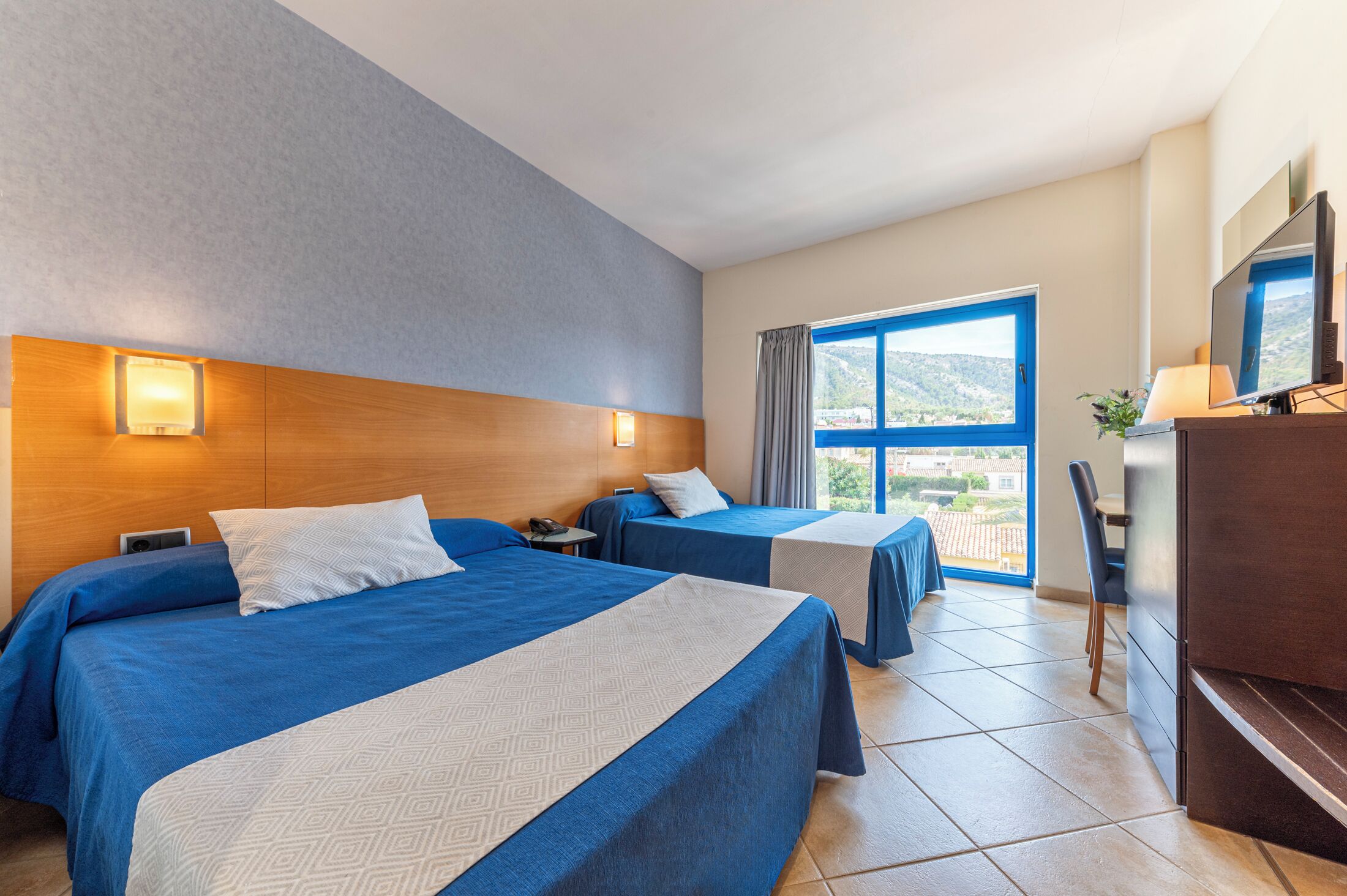 A hotel room with two beds, a desk, a chair, and a large window with a view.