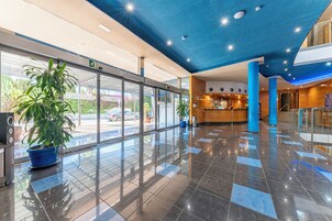 A modern hotel lobby with a glass entrance, a reception desk, and potted plants.