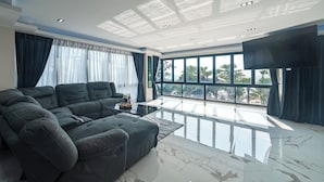 A modern living room with a large sectional sofa, a flat-screen TV, and large windows offering a view of palm trees. - Karon Sea Sands Resort (Karon)