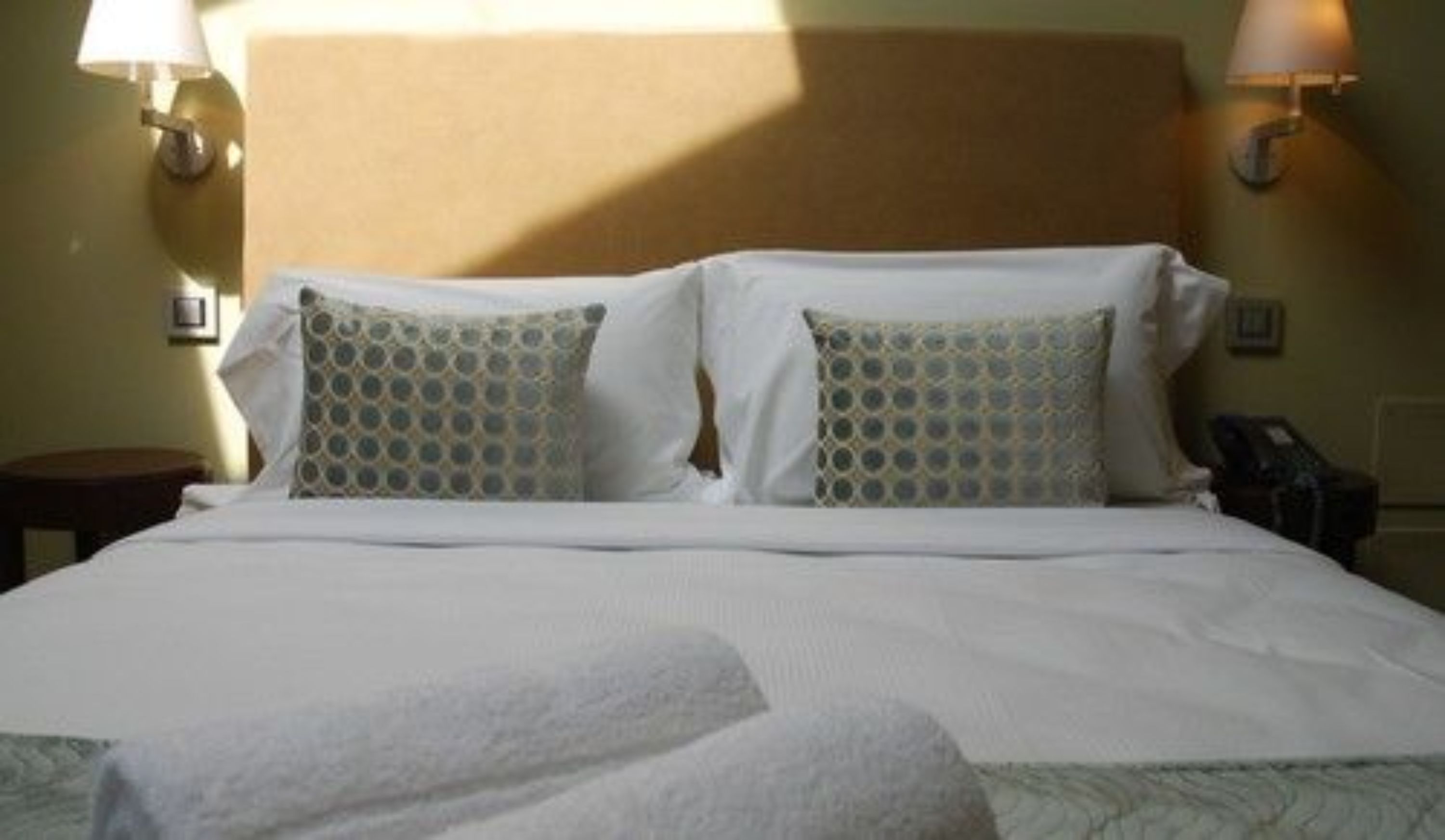 Una cama bien hecha con sábanas blancas y dos almohadas estampadas, una lámpara de noche y una mesita.