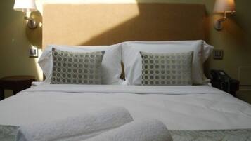 Una cama bien hecha con sábanas blancas y dos almohadas estampadas, una lámpara de noche y una mesita.