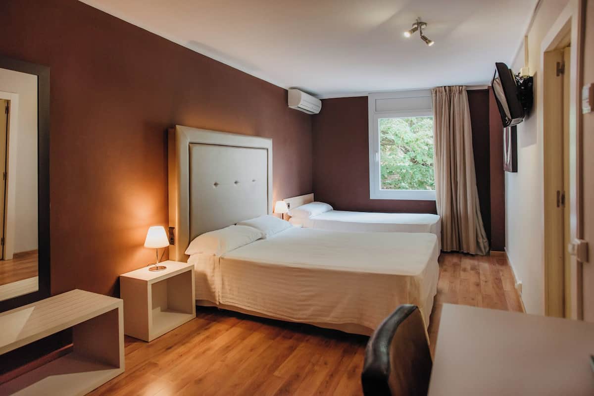 A hotel room with two beds, a desk, a chair, a nightstand with a lamp, a TV mounted on the wall, and a window with curtains.