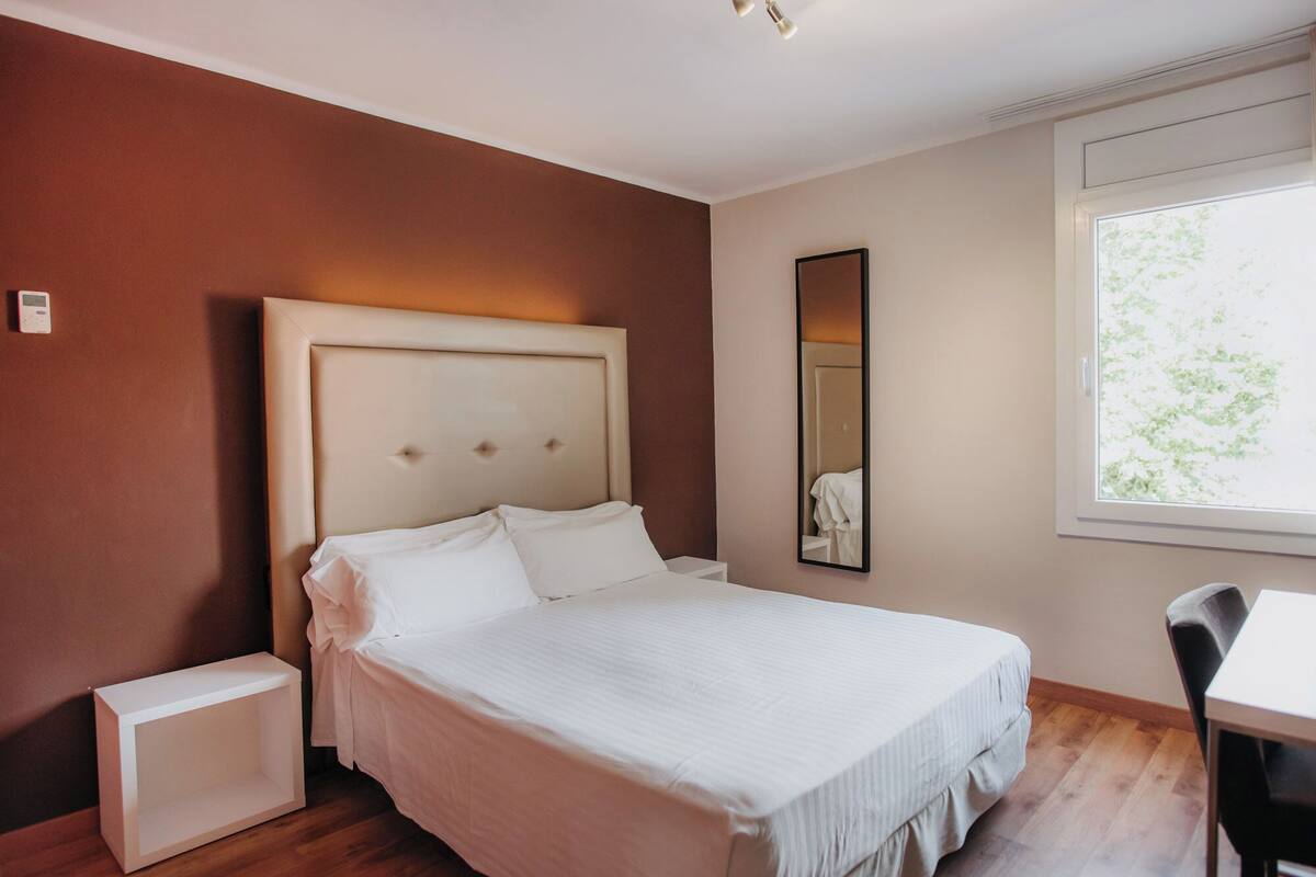 A bedroom with a bed, bedside table, and a window with a view of trees.