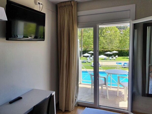 Habitación de hotel con televisión montada en la pared, un escritorio con un control remoto, y una puerta corrediza de vidrio que da a un área de piscina con sombrillas y sillas reclinables.