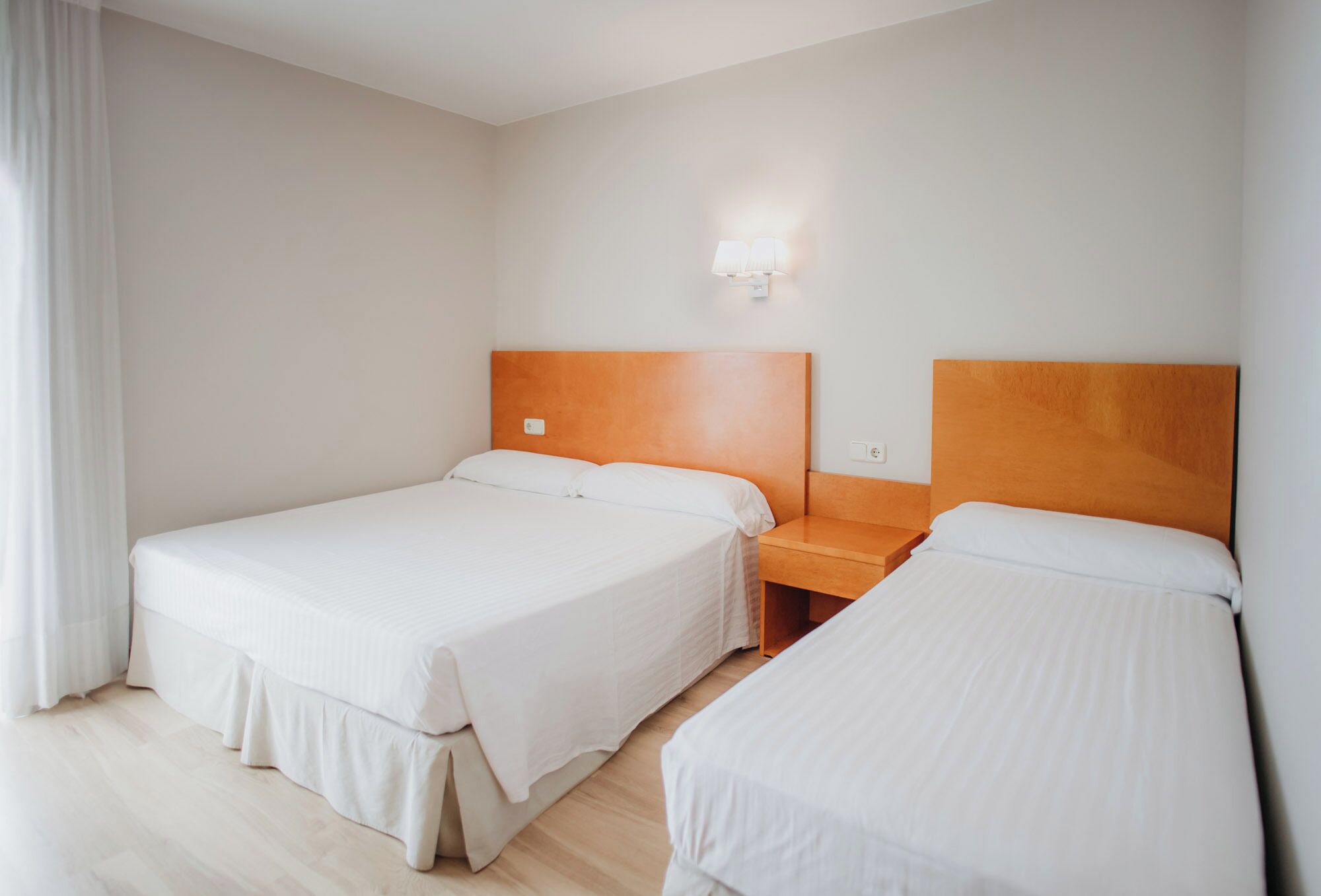 Habitación de hotel con dos camas individuales, cabecero de madera, mesita de noche y lámpara de pared.
