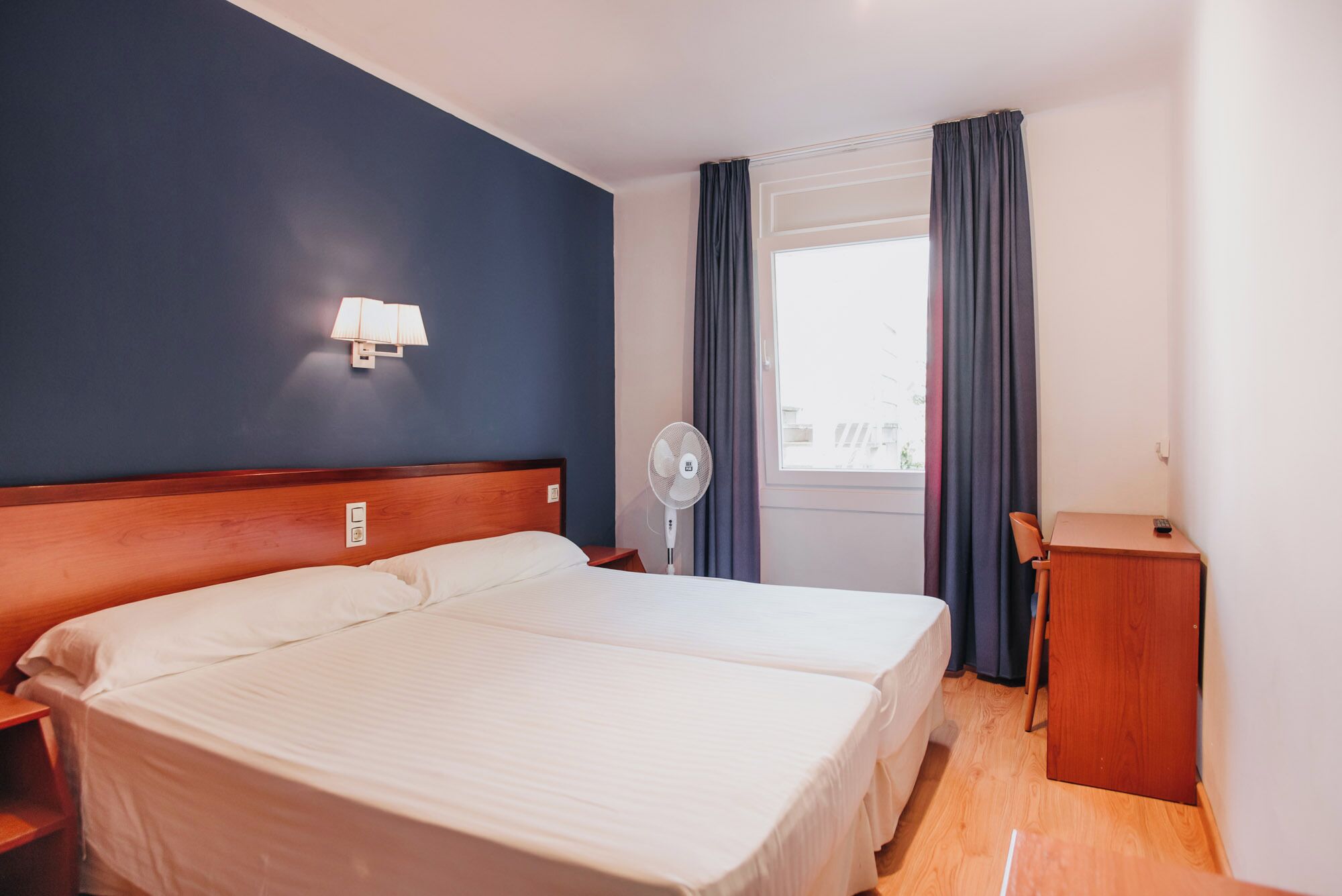 A hotel room with two beds, a bedside table, a desk, a lamp, a fan, and a window with curtains.
