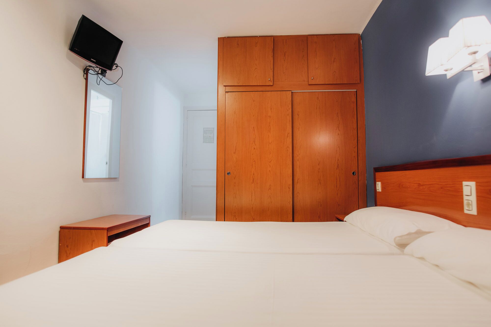 Une chambre avec une armoire en bois, un lit avec une literie blanche, une petite table de chevet, un téléviseur fixé au mur et une fenêtre laissant entrer la lumière naturelle.