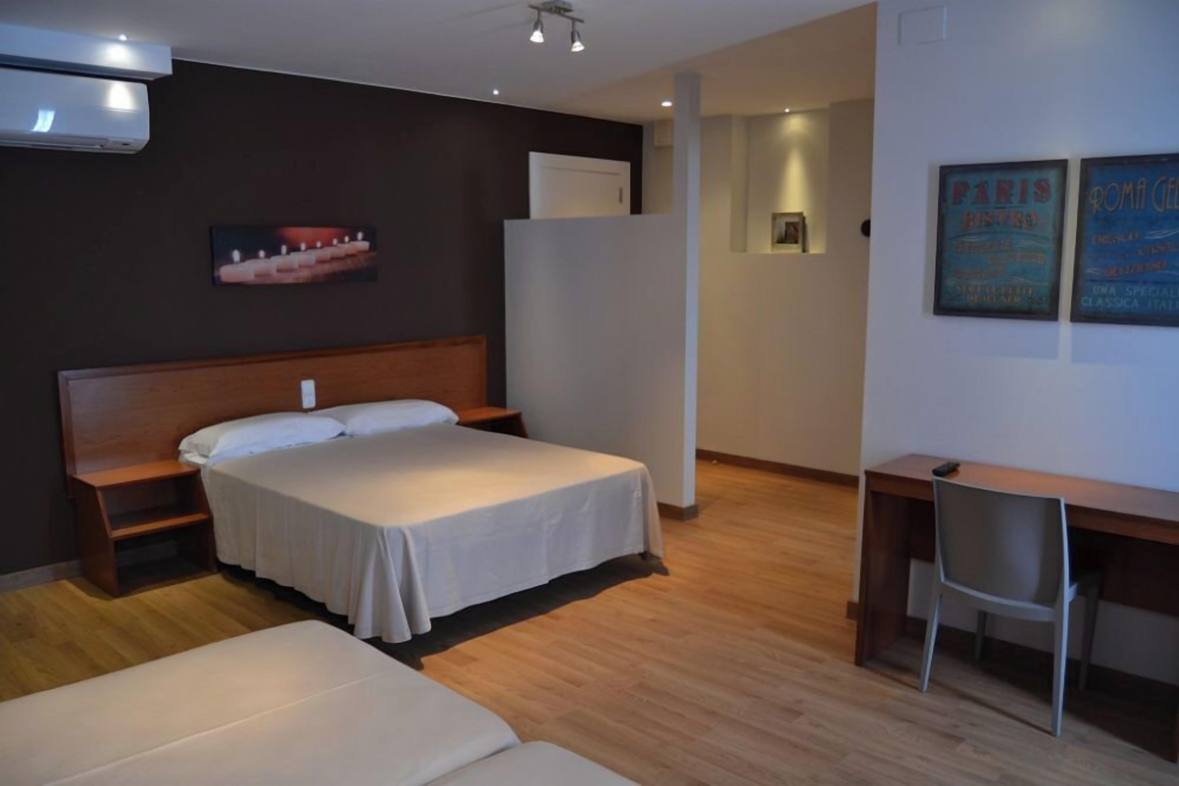 A hotel room with a bed, a desk, and a chair. There is a picture on the wall and an air conditioner above the bed.