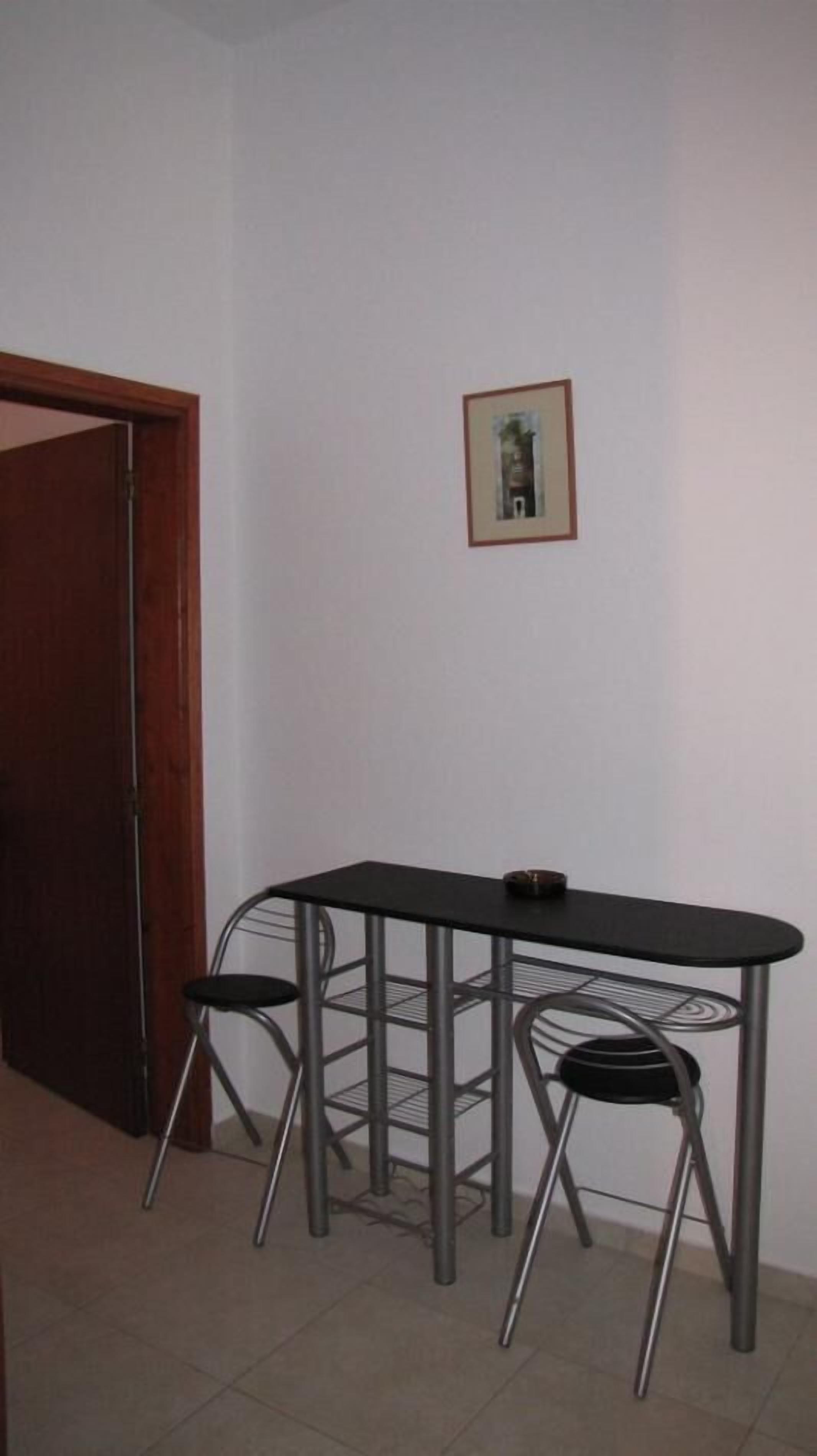 Een kleine kamer met een houten deur, een zwart metalen tafeltje met twee stoelen, en een lijst met een afbeelding aan de muur.