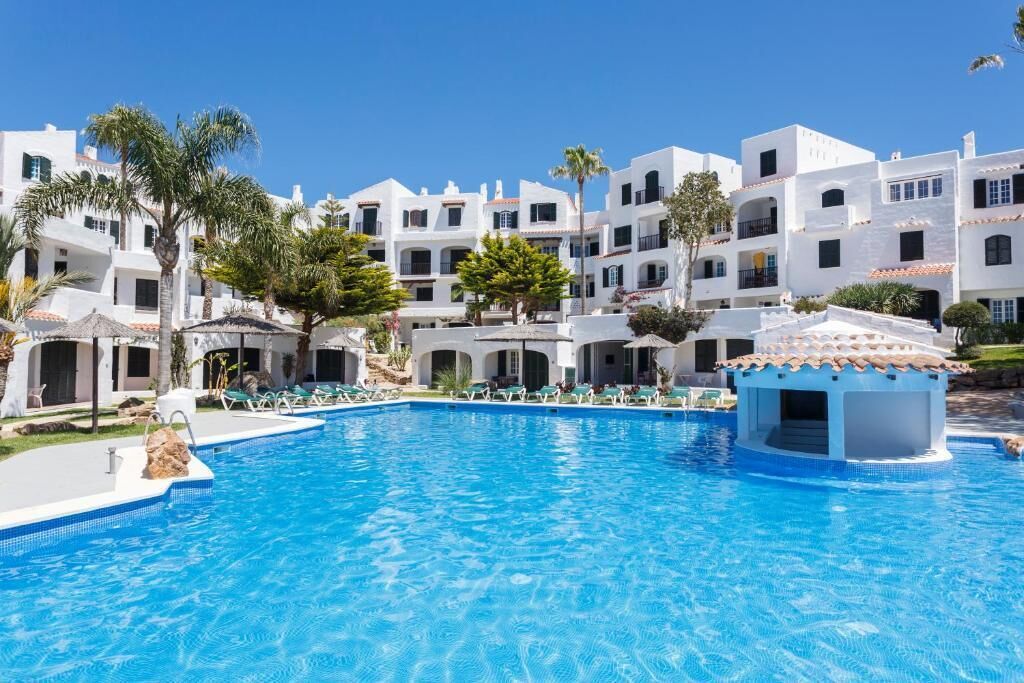 Une piscine entourée de bâtiments blancs dotés de balcons, de palmiers et surplombée d’un ciel bleu azur.